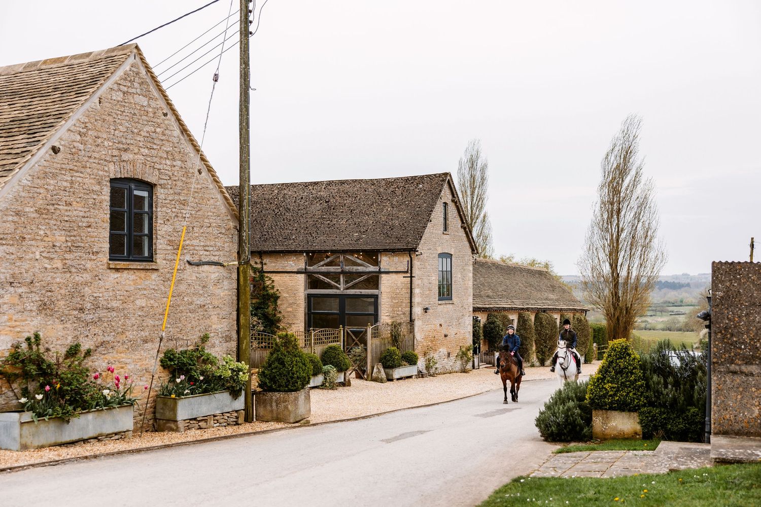 Stone buildings line a curved rural road with trees and greenery in the English countryside.