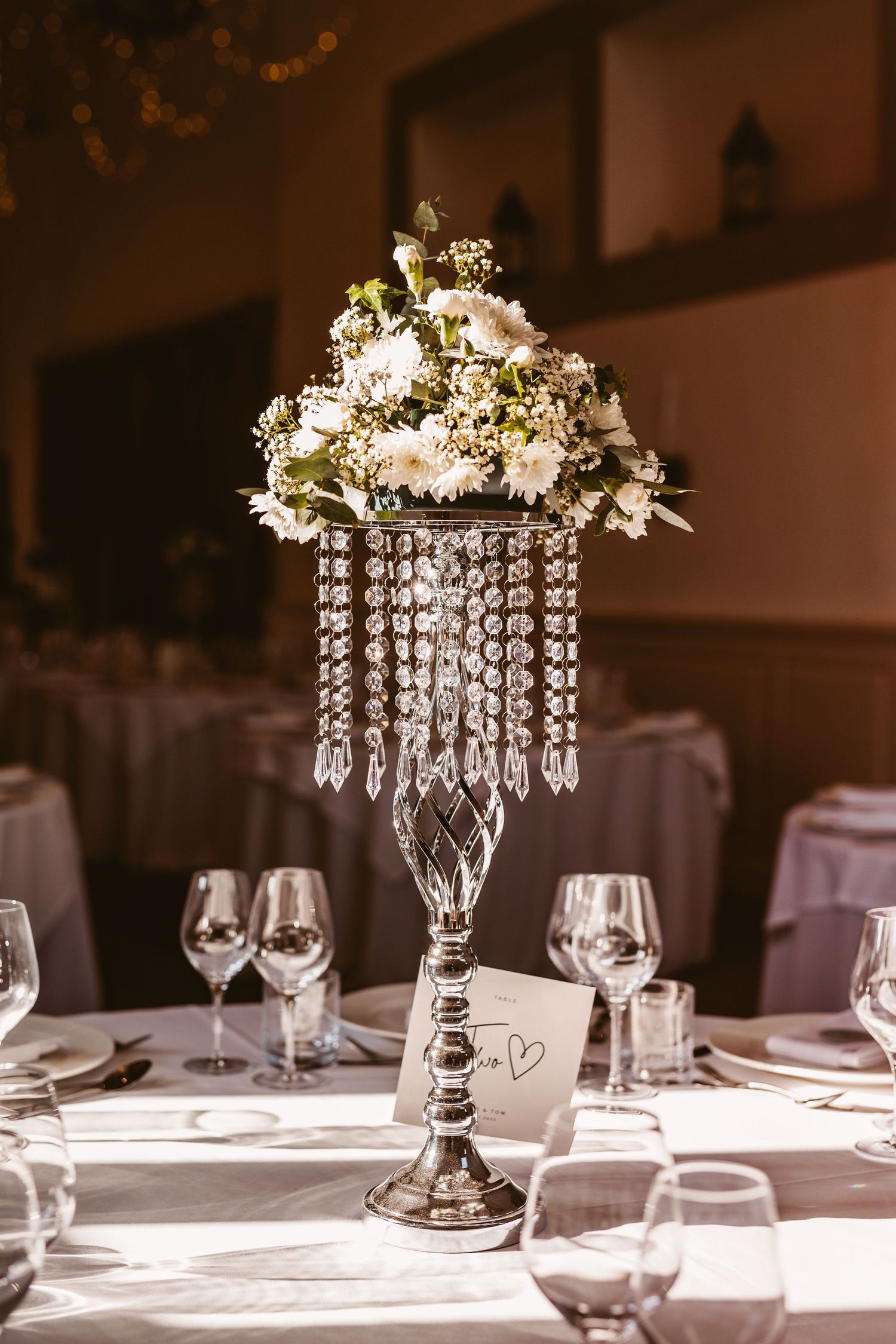Crystal chandelier centerpiece with white flowers adorns an elegant wedding reception table setting in warm sunlight.