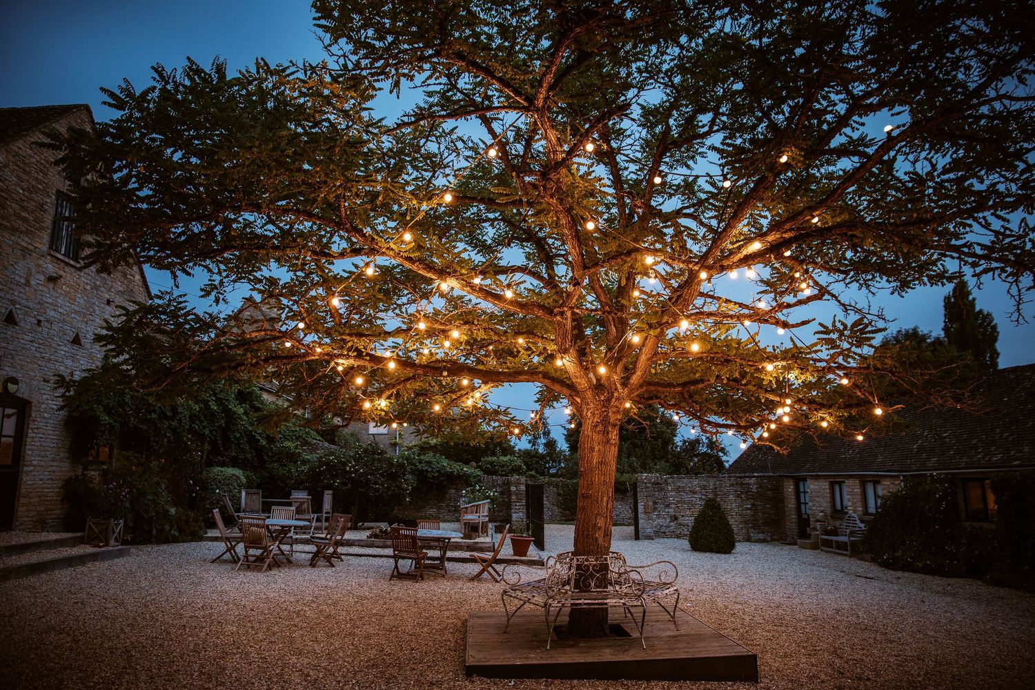String lights illuminate a large tree over a gravel patio with bistro tables and chairs at dusk.