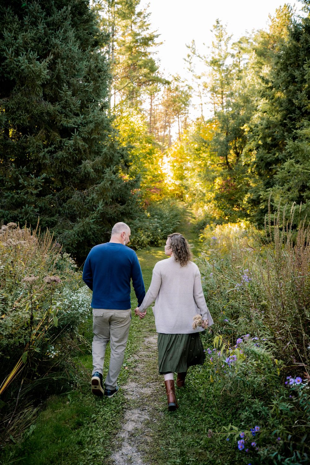 A couple walks hand in hand down a scenic nature path surrounded by lush greenery and wildflowers at sunset.