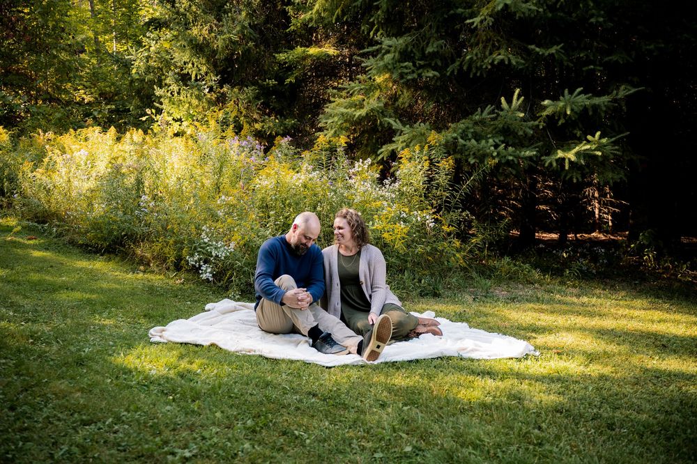 A couple enjoys a romantic picnic on a white blanket in a sunlit grassy meadow surrounded by trees.