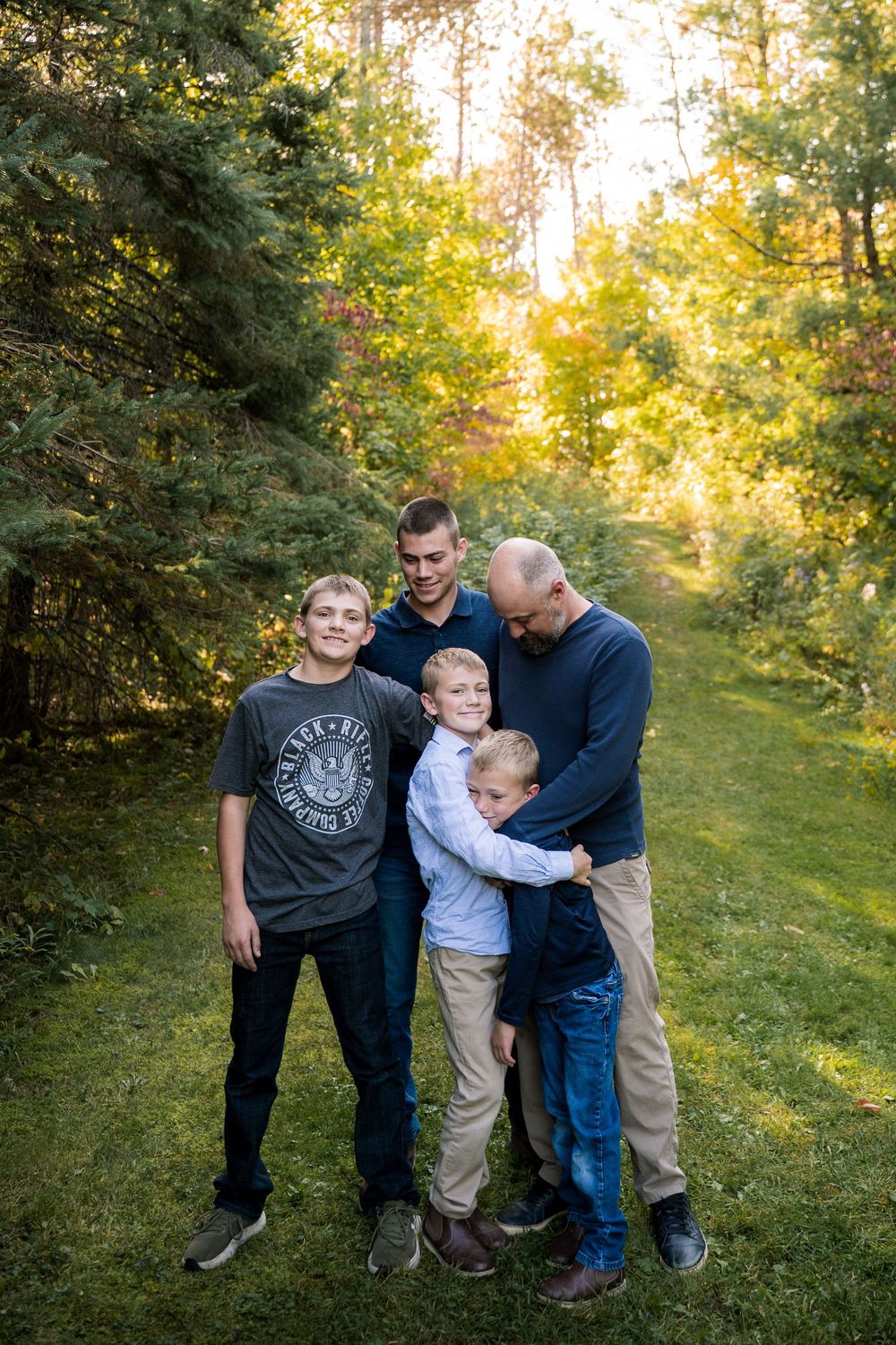 Family members huddle close together for a casual outdoor portrait in a sunlit wooded area.