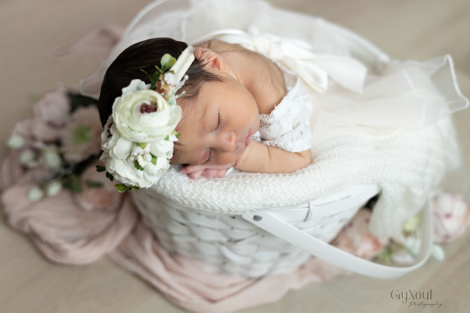 A sleeping newborn baby wrapped in white lace fabric lies peacefully on soft textured bedding.