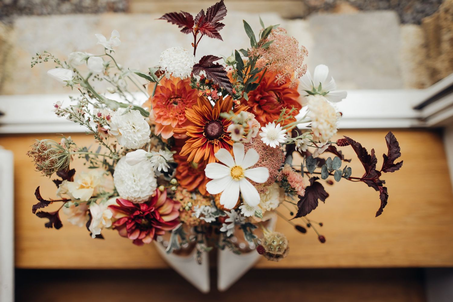 Stunning autumn wedding bouquet featuring orange dahlias, white cosmos, and burgundy foliage on wooden surface.