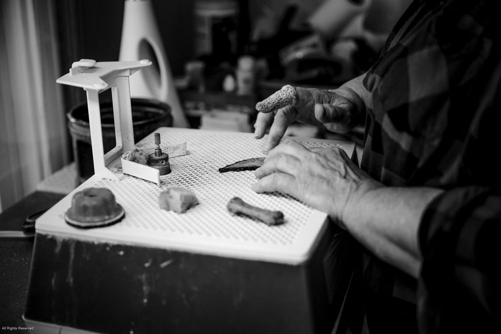 Black and white image showing hands working at a desk with various tools and a small lamp.