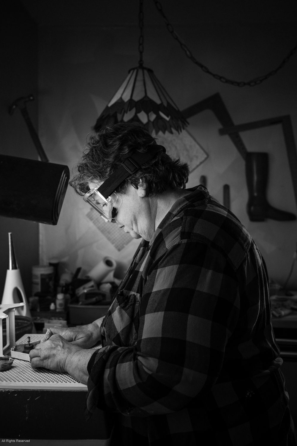 Black and white photo showing someone in a plaid shirt working at a desk under a hanging pendant lamp.