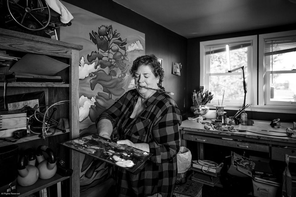 A black and white photograph shows an artist in a plaid shirt standing in a studio workspace with artwork and supplies.