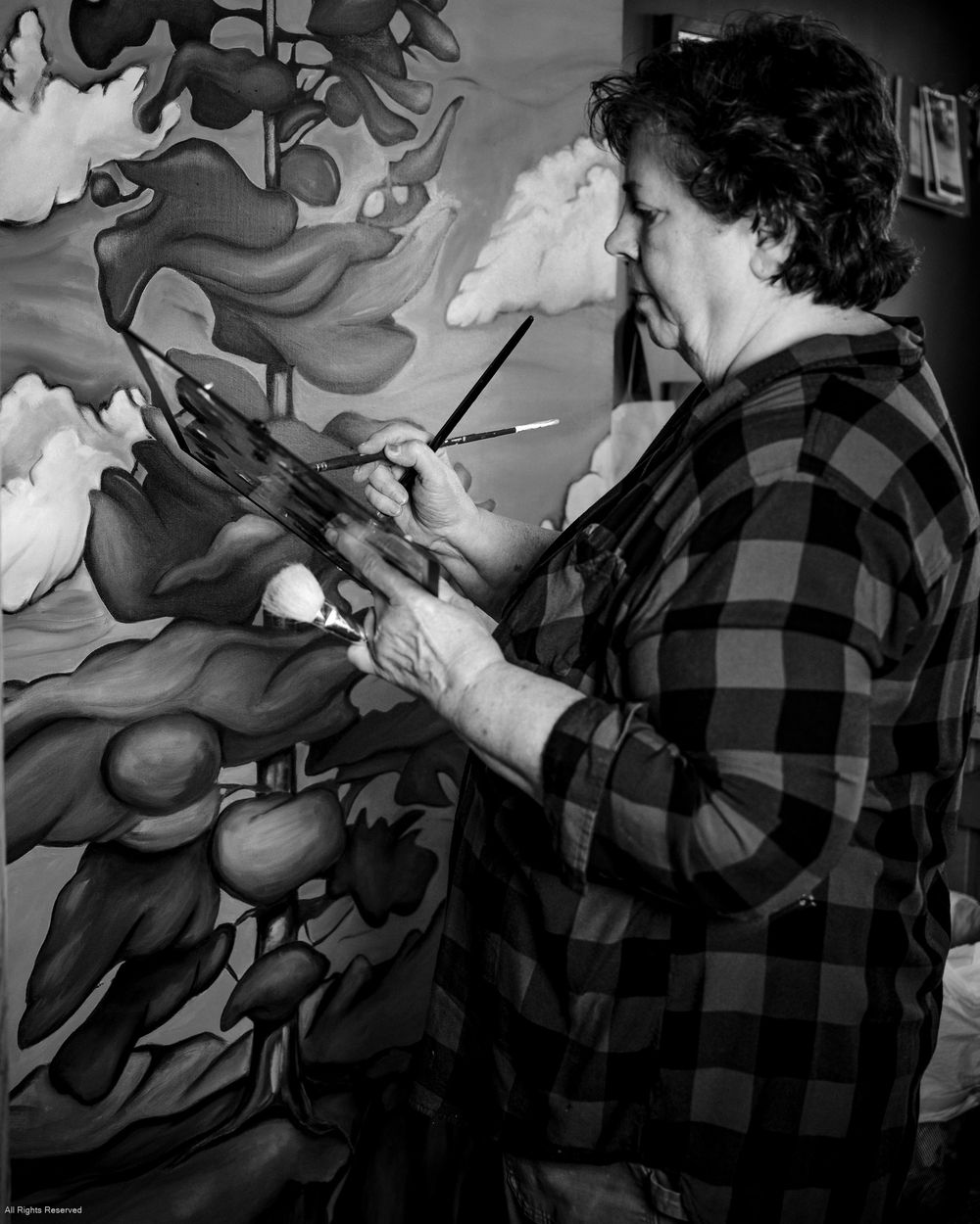 Artist in plaid flannel shirt works on a vibrant painting while holding a palette in a black and white photograph.