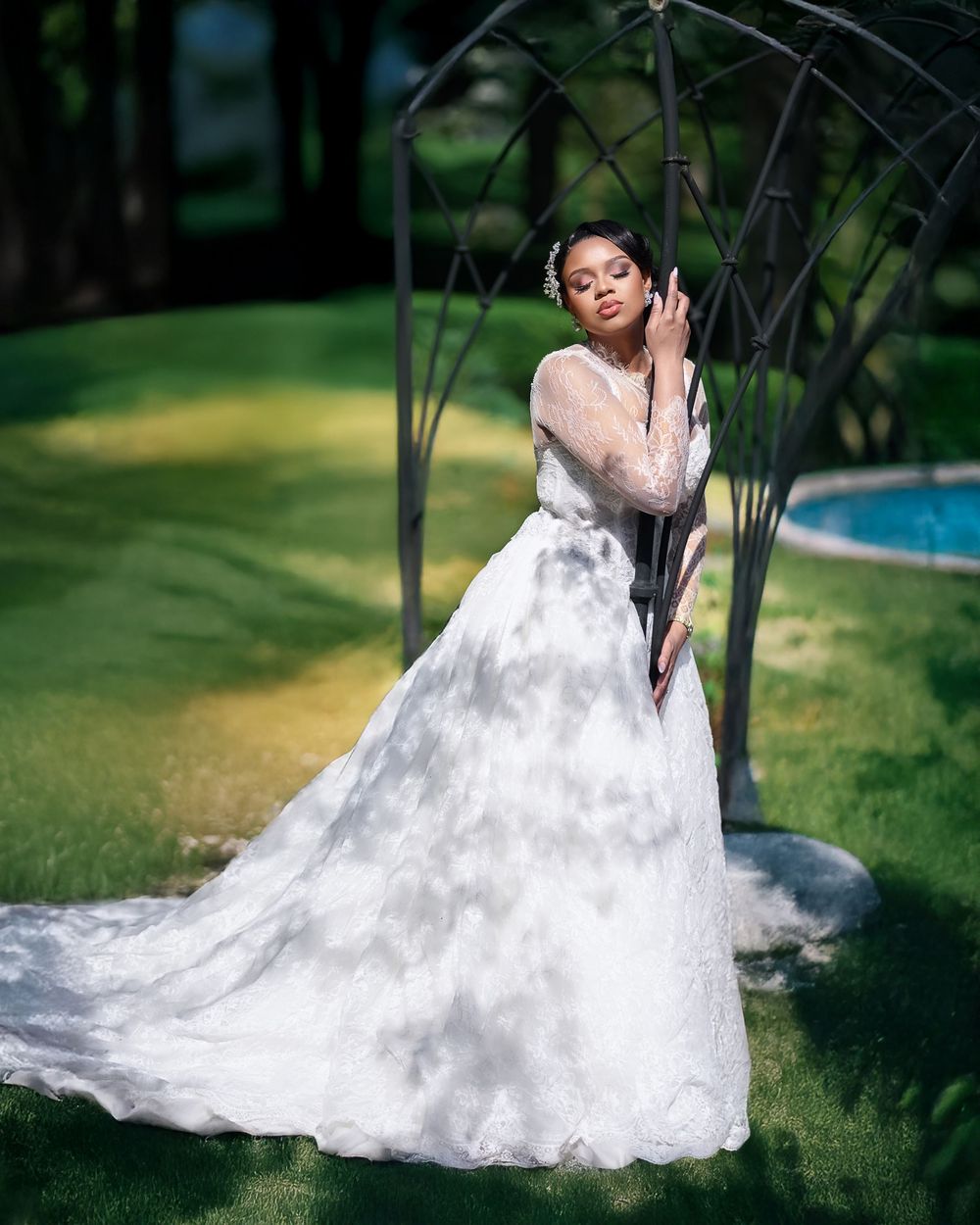 A bride in a flowing white lace gown poses gracefully in an outdoor garden setting with greenery and a pool in background.