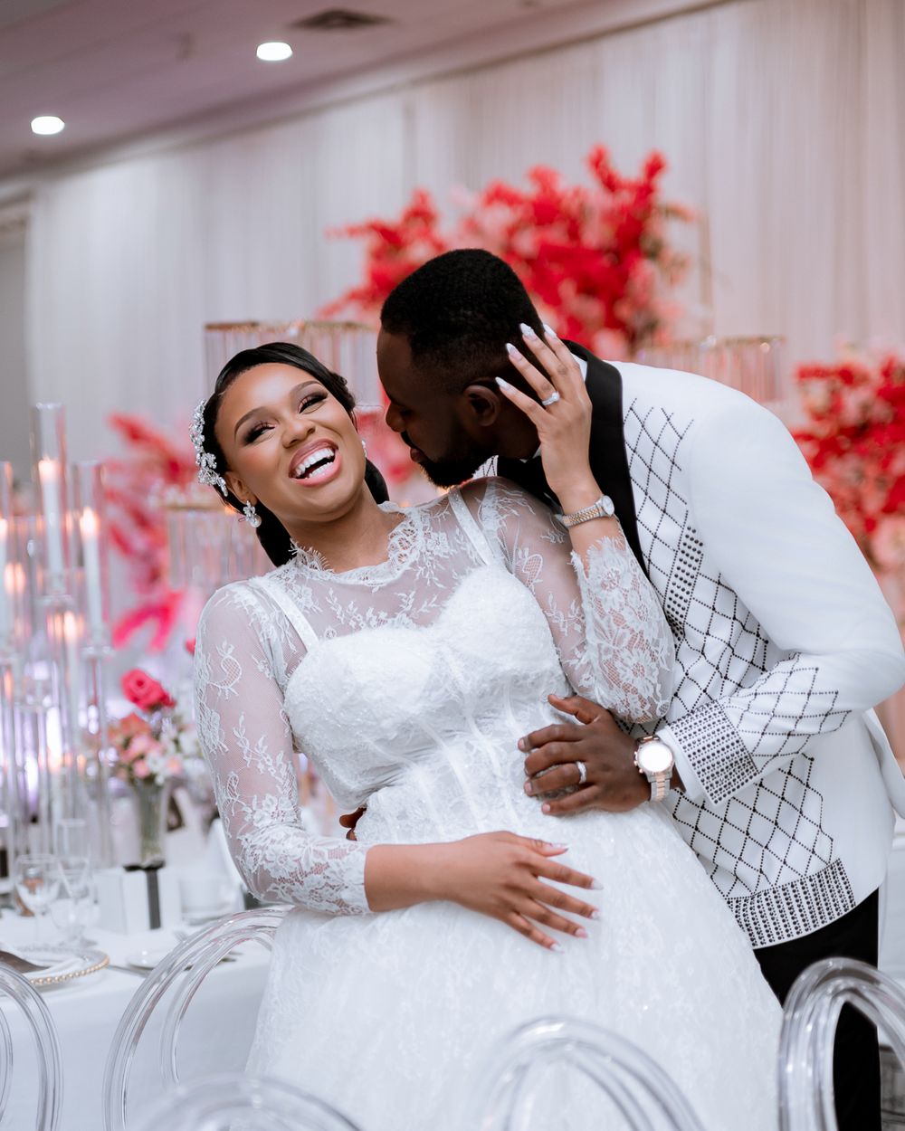 A couple shares a romantic moment in their wedding attire with red florals and crystal decor in the background.