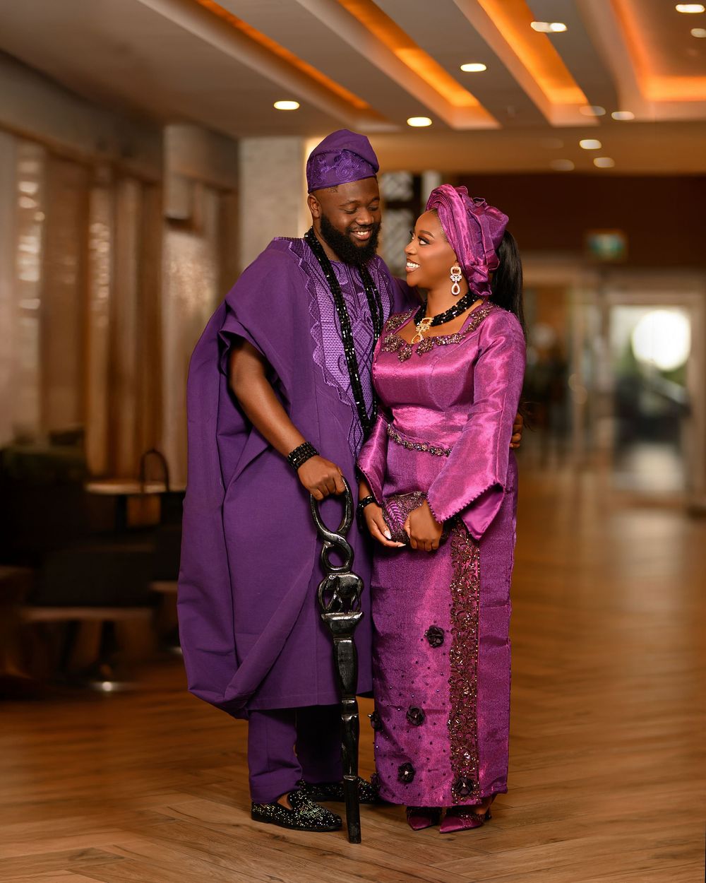 A couple wearing coordinated purple and pink traditional African attire poses together in an elegant indoor setting.