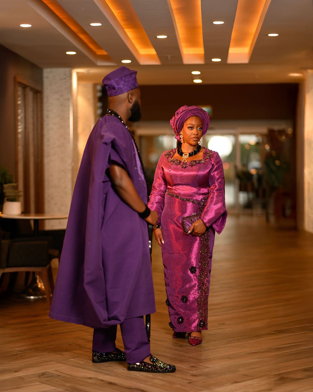 Two people in vibrant purple and pink traditional clothing interact in a warmly lit hallway.