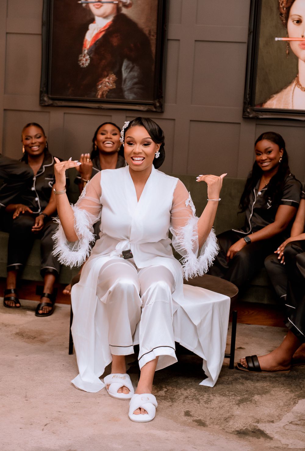 Joyful bridal party scene with person in white feathered robe sitting on chair while others laugh in background.