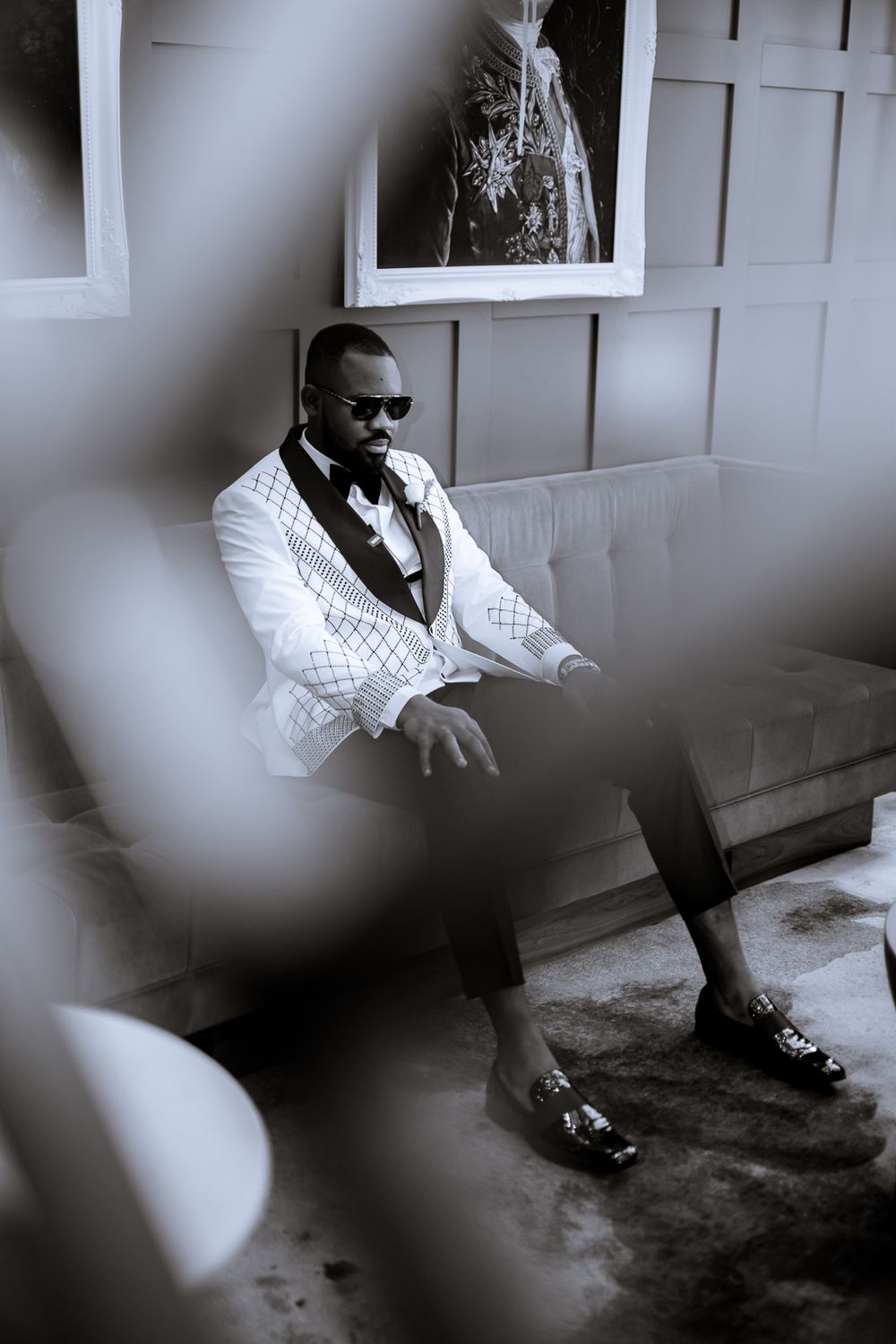 A stylish person wearing a white suit jacket, dark sunglasses and loafers sits in a pensive pose by a window.