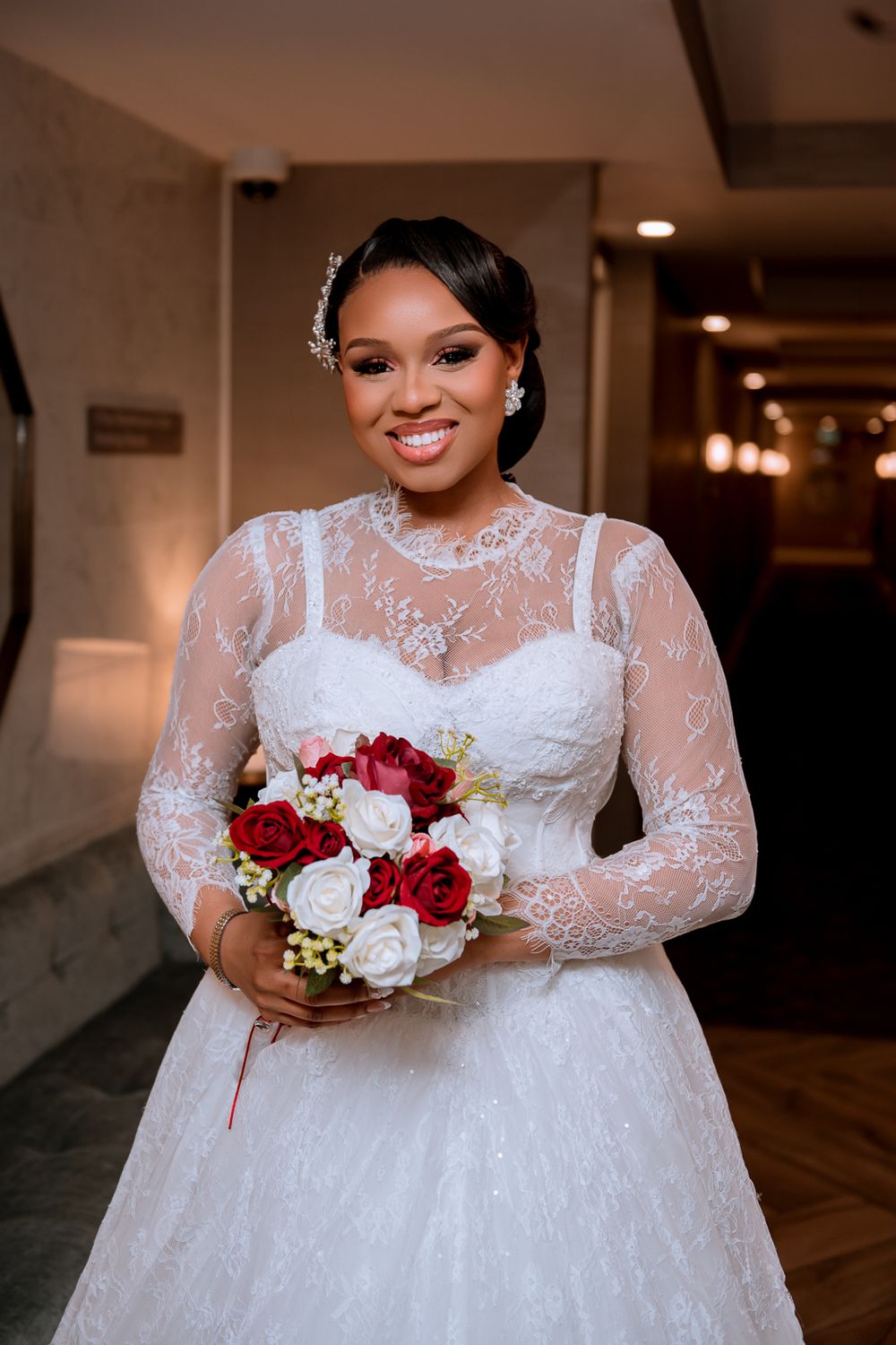 Beautiful bride in lace long-sleeve wedding gown holds red and white rose bouquet in indoor hallway.