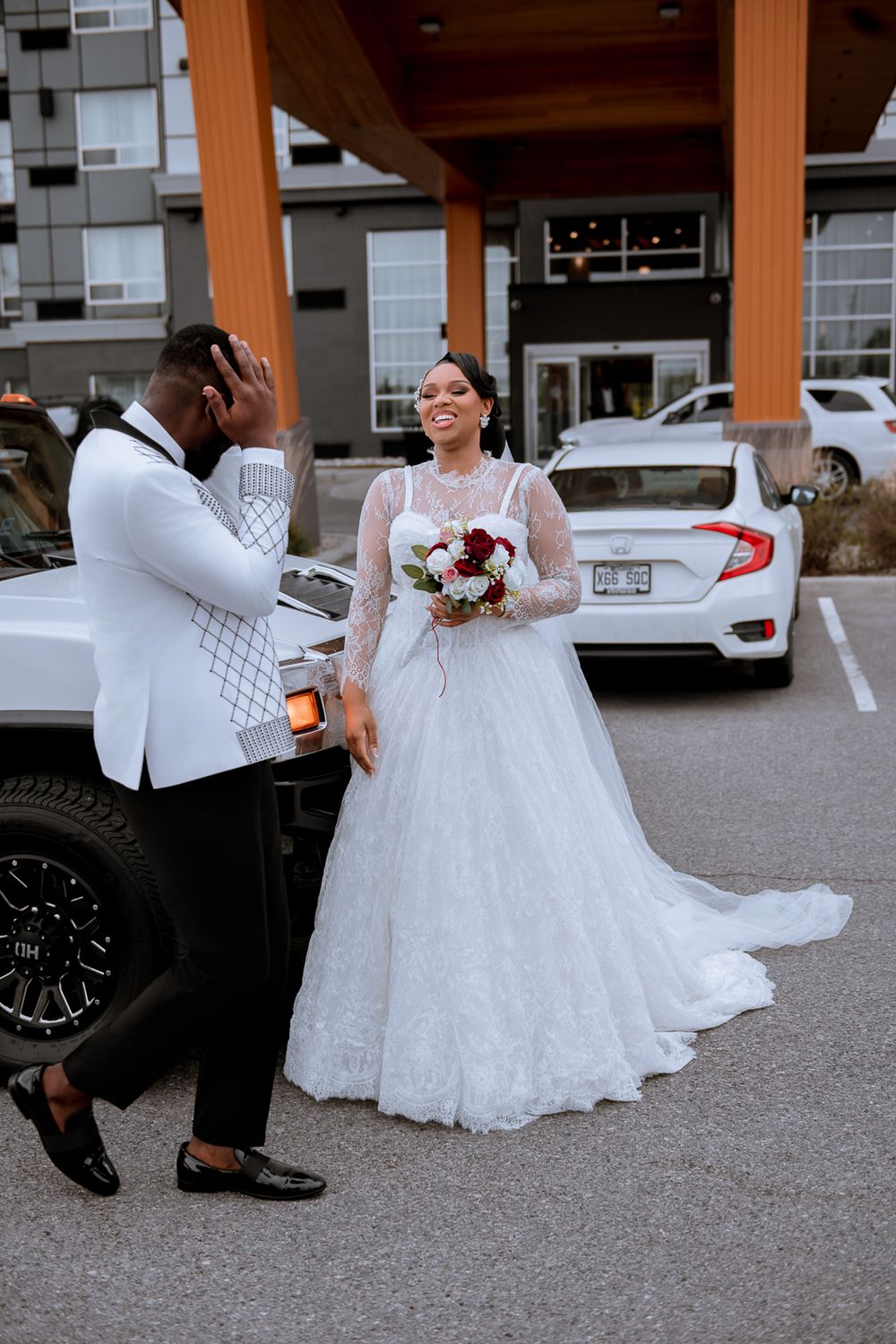 A bride in a white lace wedding gown and groom in a white jacket react emotionally in a parking lot.