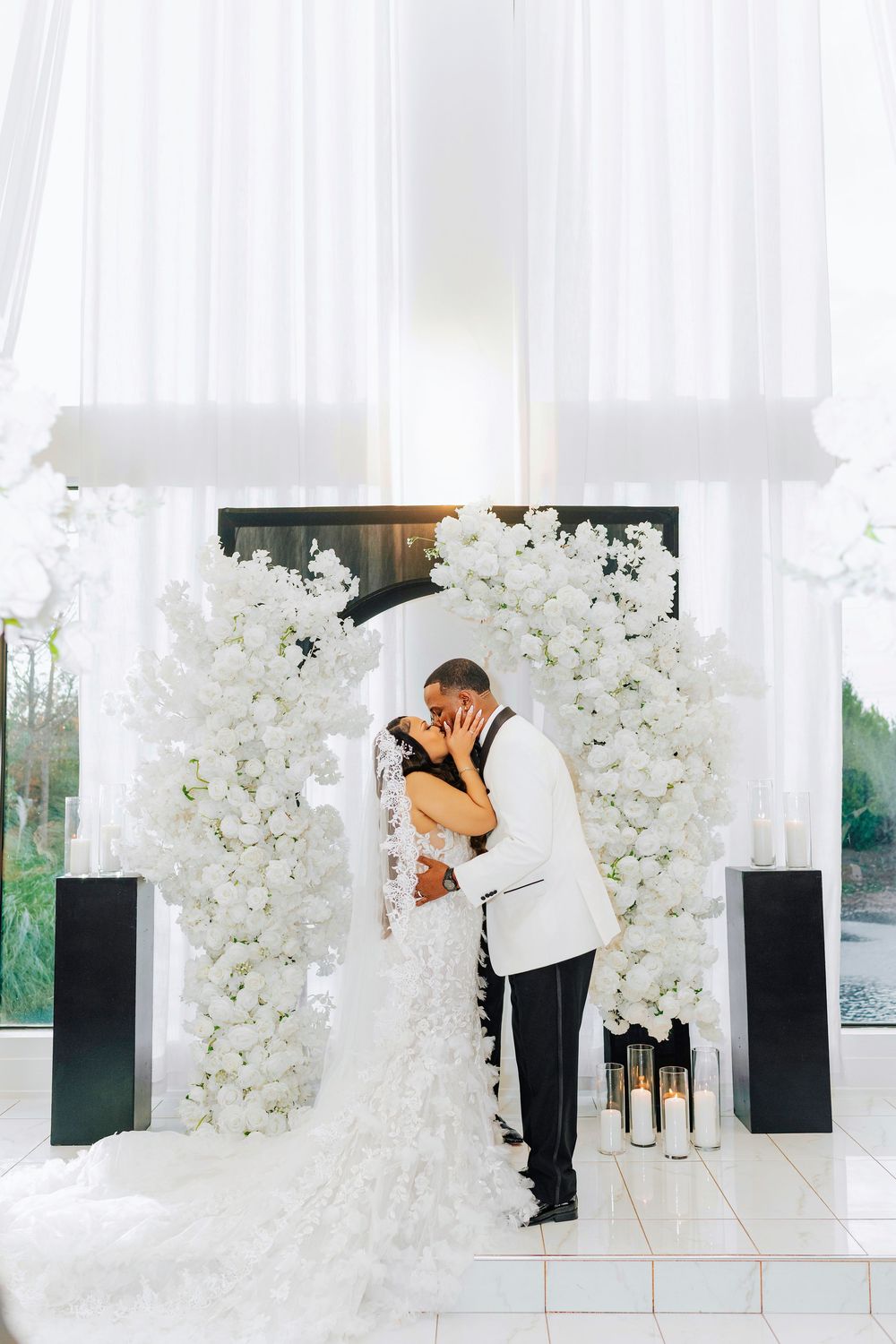 Elegant wedding ceremony setup with white floral arrangements and sheer curtains in a modern indoor venue.