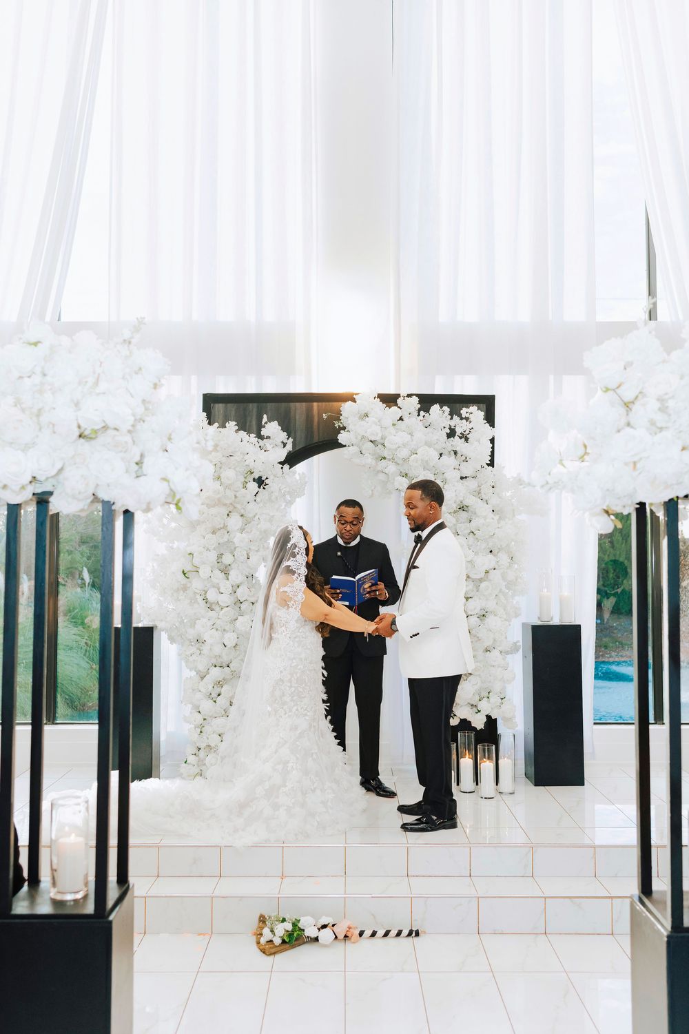 Wedding couple embracing in an all-white modern venue decorated with luxurious floral arrangements and floor-to-ceiling windows.