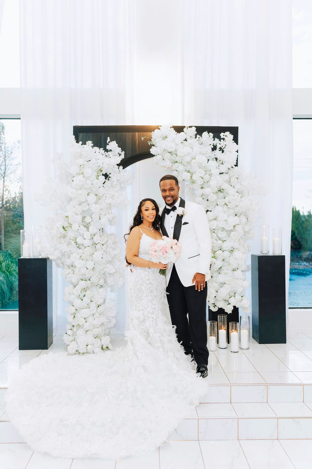 Elegant wedding ceremony setup with white floral arrangements on black pedestals and a waterfront view in the background.