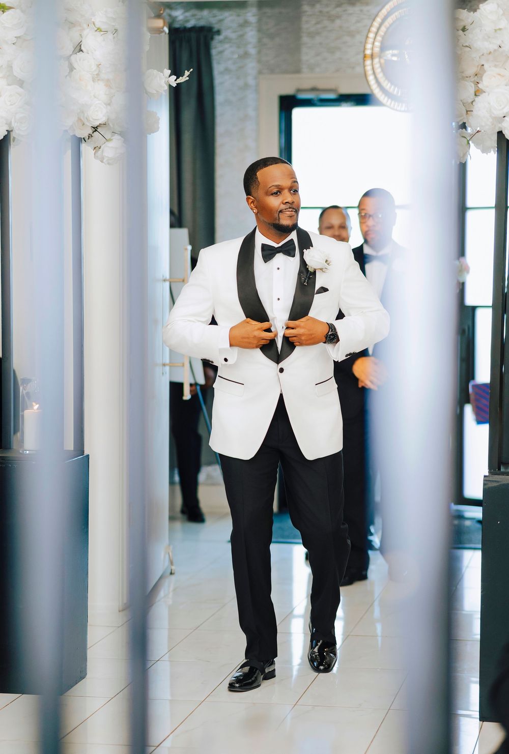 A groom in a crisp white tuxedo jacket and black bow tie walks confidently through an elegant hallway with white columns.