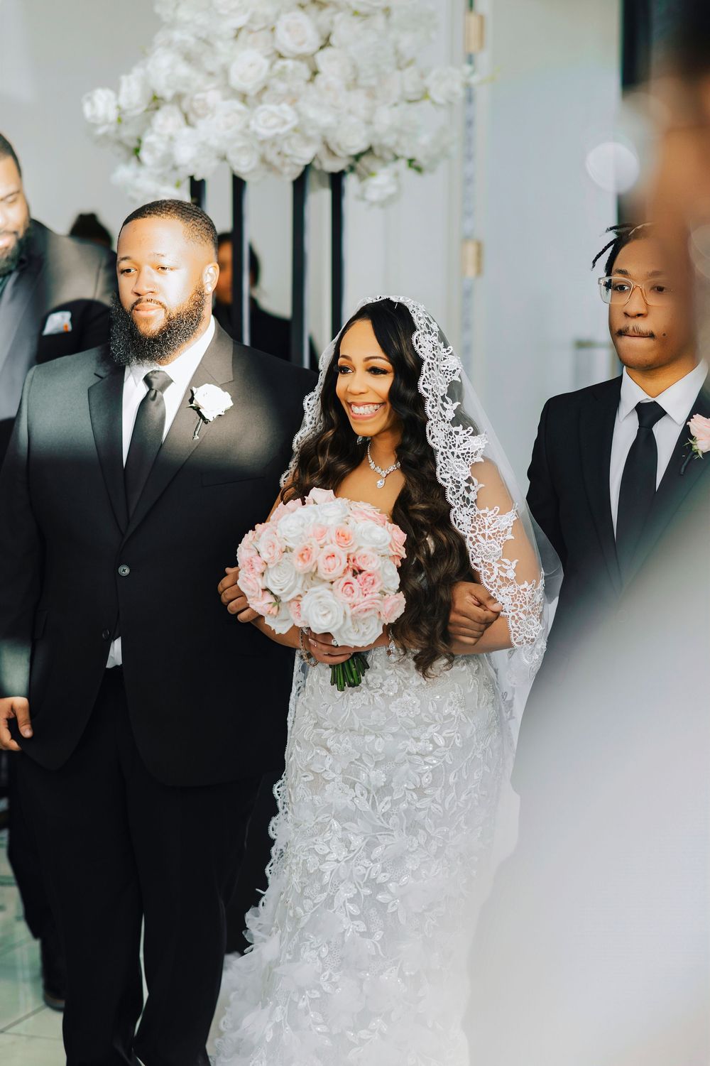 Wedding party in black suits and white lace dress walk together holding pink and white floral bouquets.