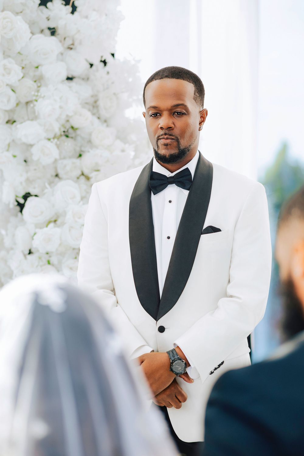 Groom wearing a classic white tuxedo jacket with black lapels and bowtie stands at wedding ceremony.