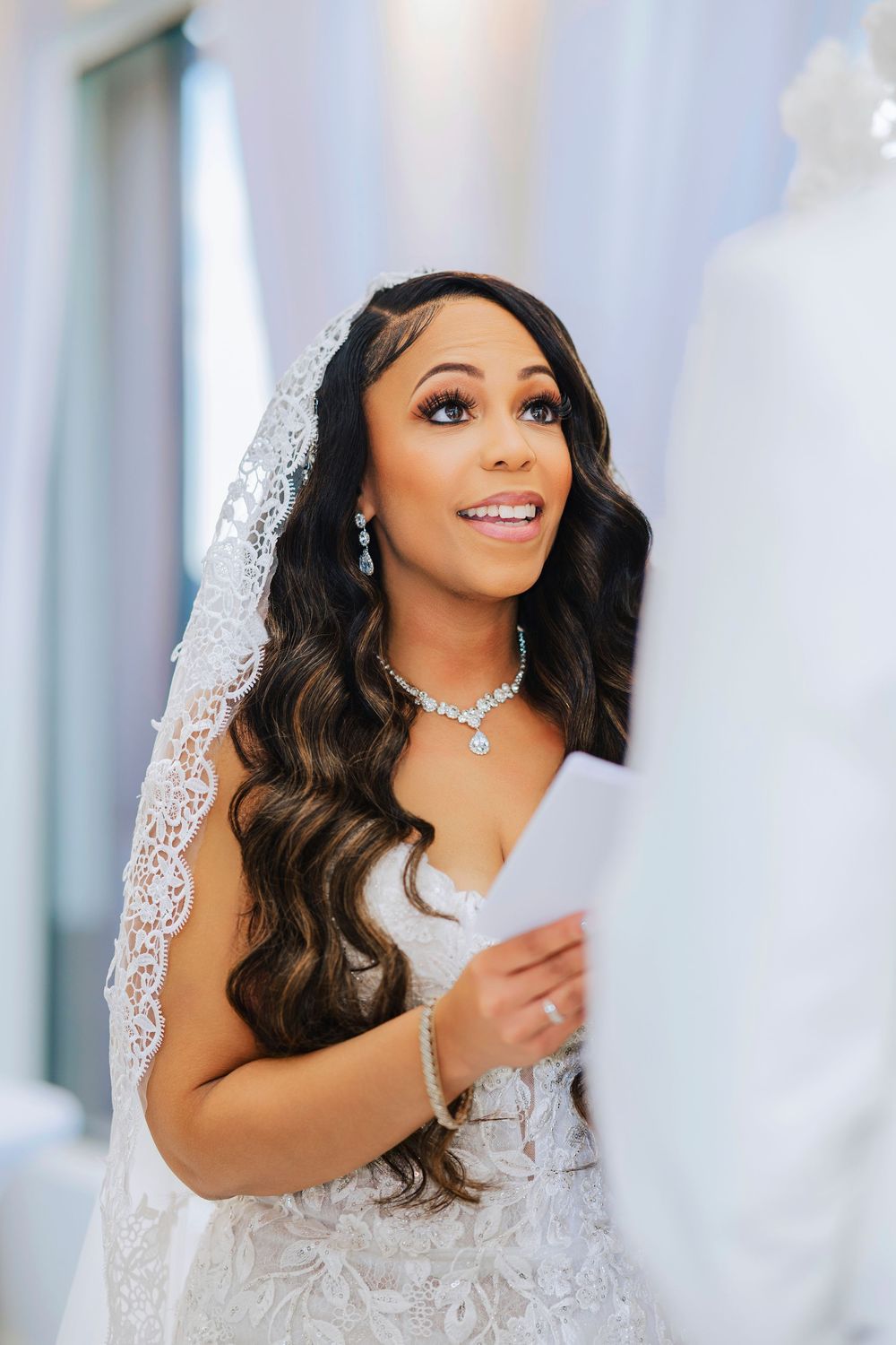 Beautiful bride in lace wedding gown with long wavy hair and crystal jewelry smiles while holding paper.