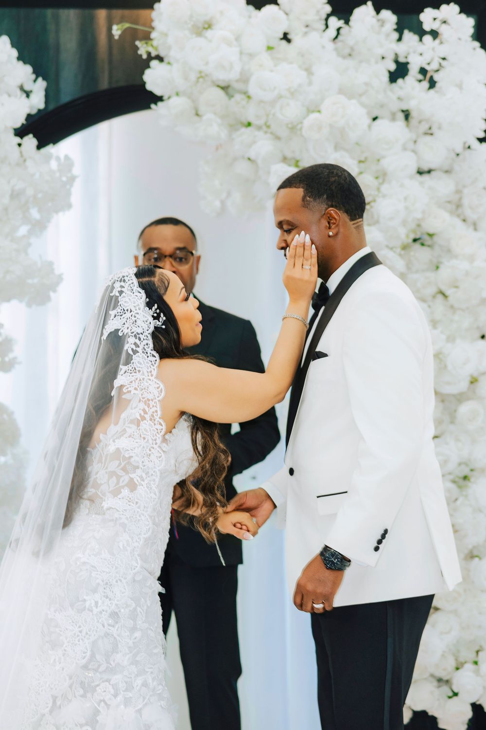 A romantic wedding ceremony moment with a bride in a lace veil and groom sharing their first kiss under white floral decor.