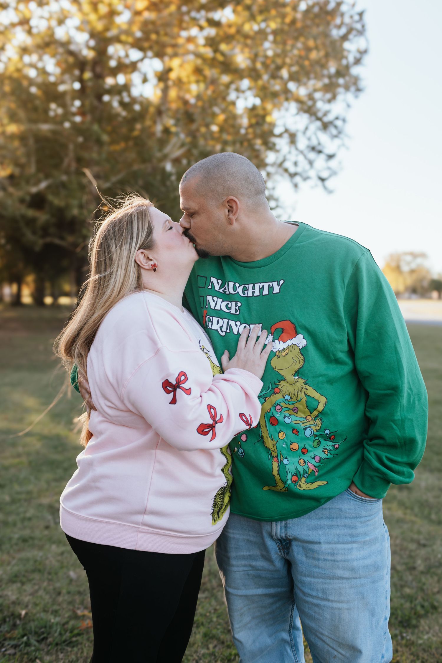 A couple wearing coordinating Christmas sweaters share a sweet moment together outdoors in a park setting.