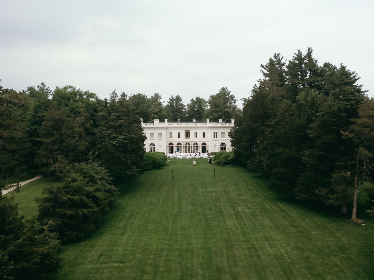 Aerial view of a grand white mansion with classical architecture set on a manicured green lawn surrounded by tall trees.
