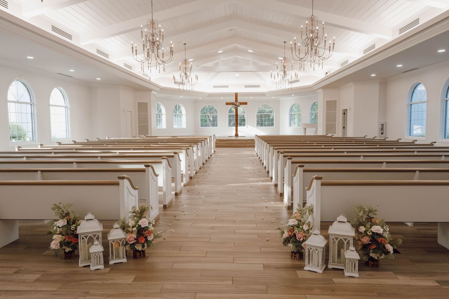 Elegant white wedding chapel interior with crystal chandeliers, arched windows, and rows of white pews.