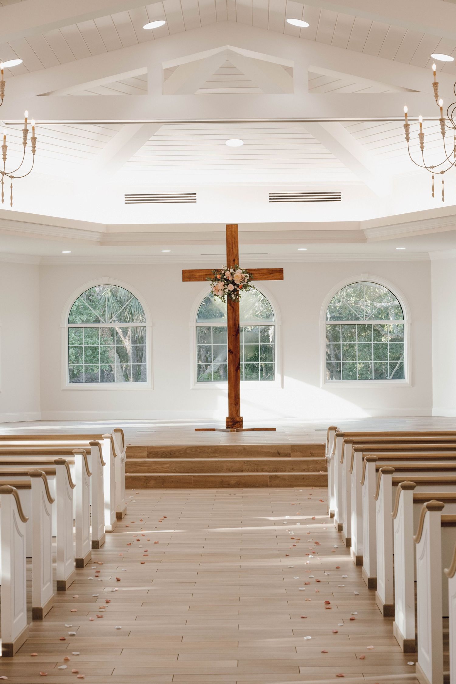 Bright white chapel interior with wooden pews, arched windows, and a large wooden cross at the altar.