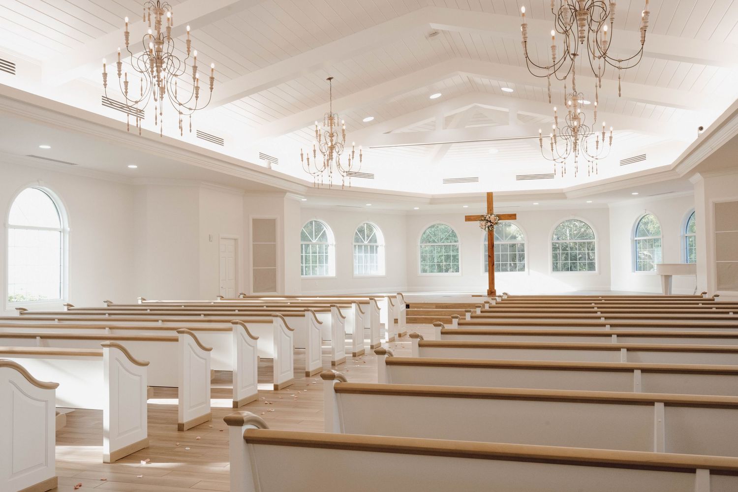 Bright white chapel interior with crystal chandeliers, wooden pews and arched windows lining the walls.