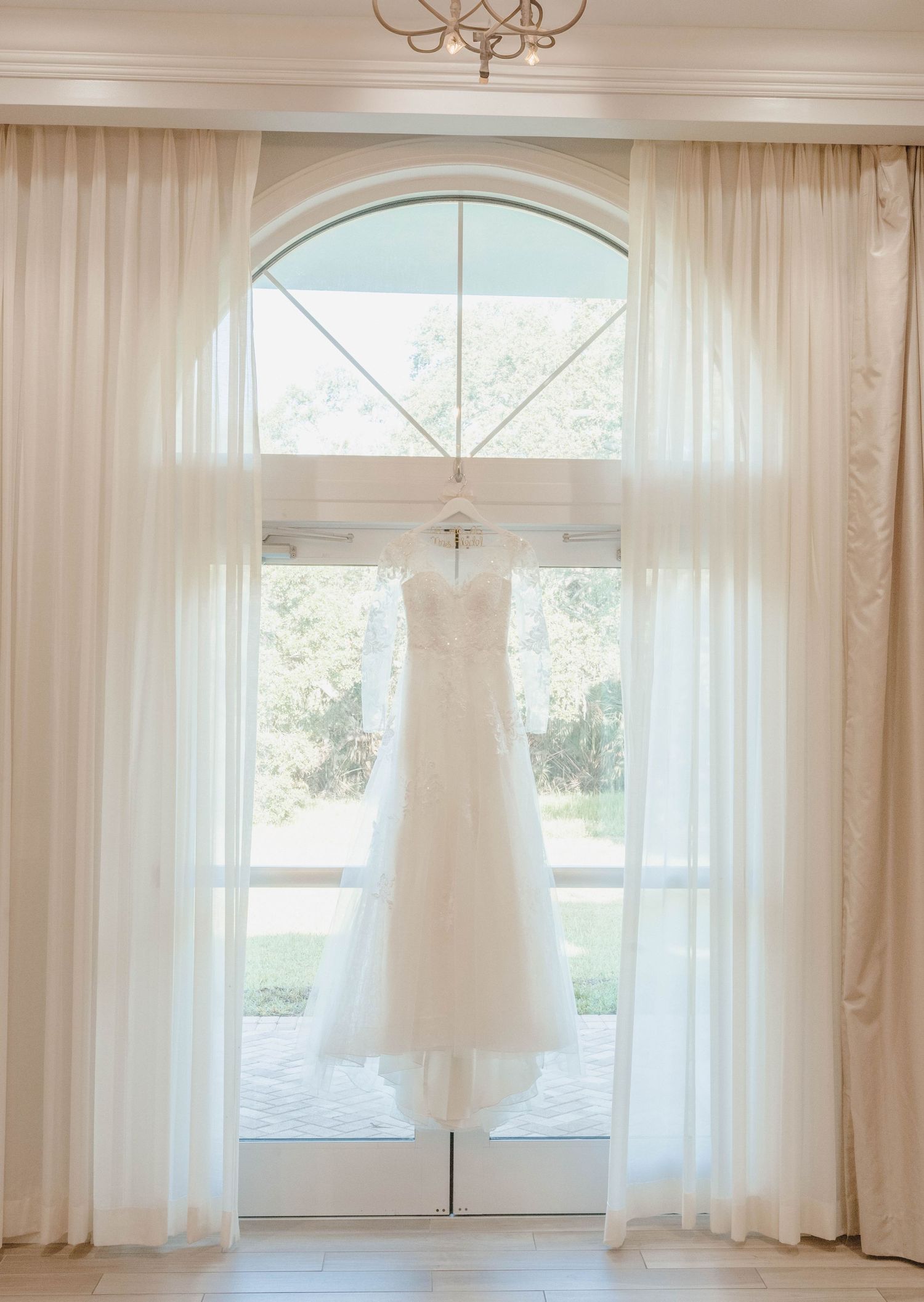 An elegant white wedding dress hangs in front of a large arched window with flowing curtains.