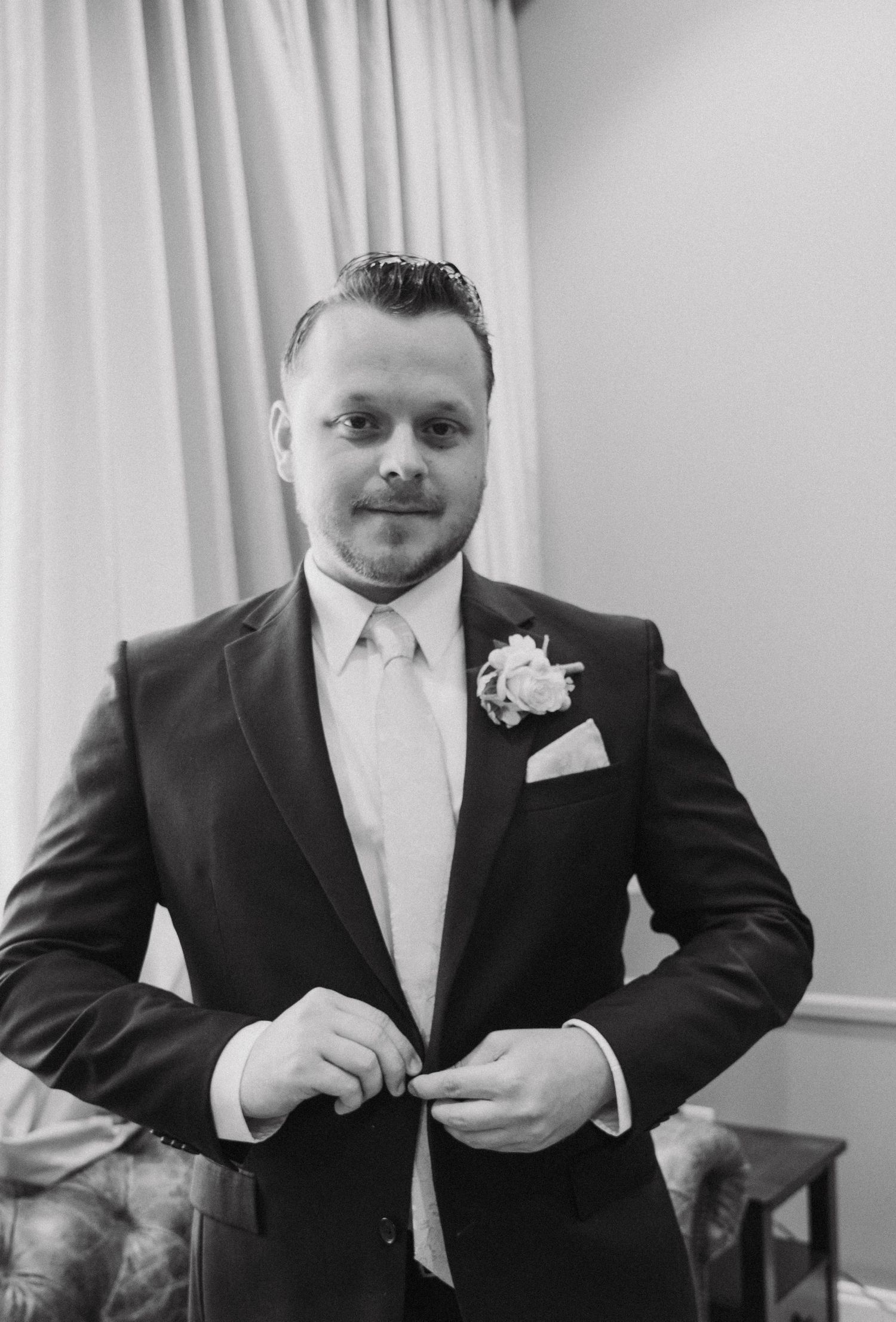 A man in a formal black suit adjusts his tie while getting ready for a wedding.