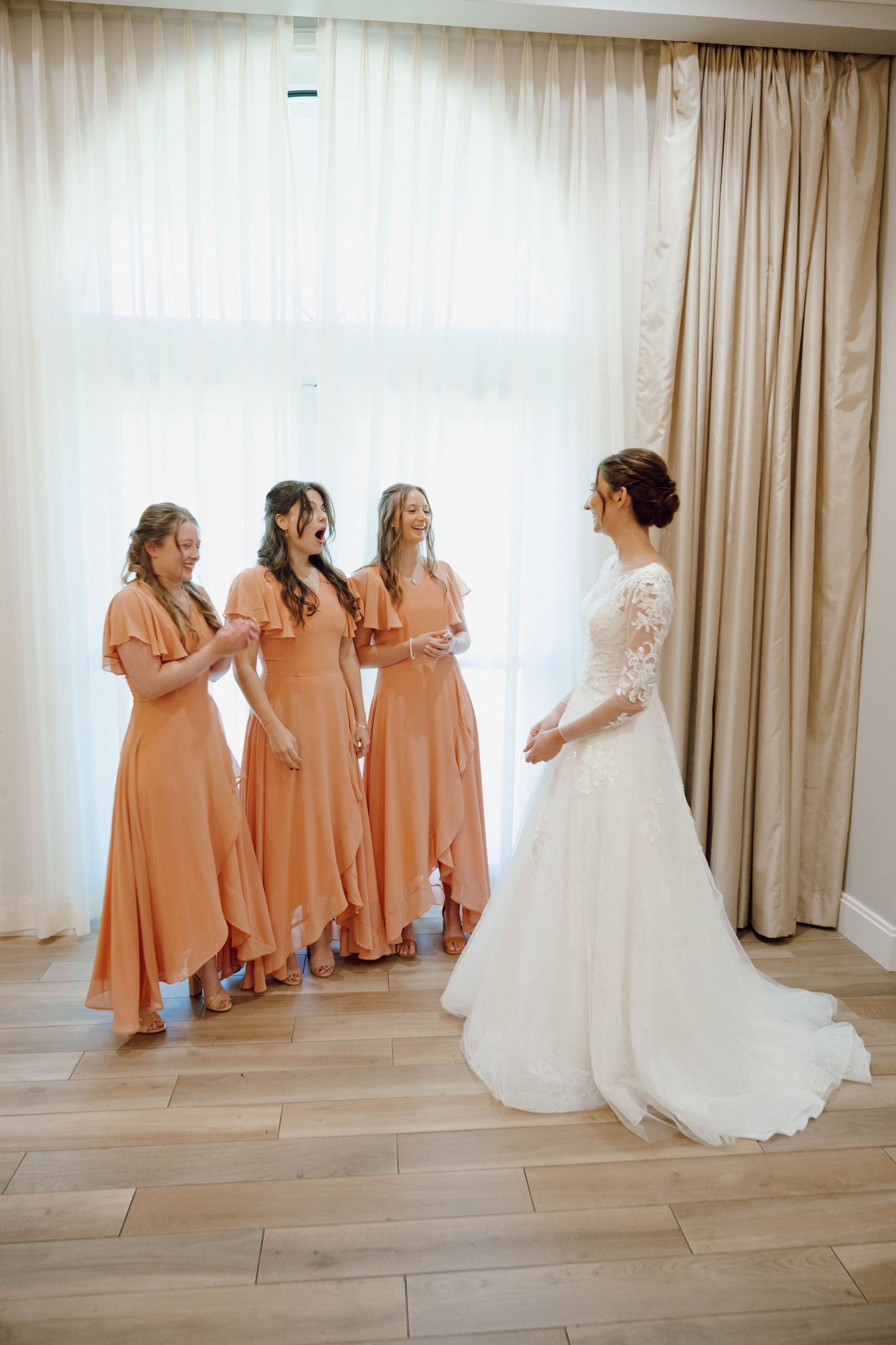A bride in a white gown stands with bridesmaids wearing peach-colored dresses in a light-filled room.