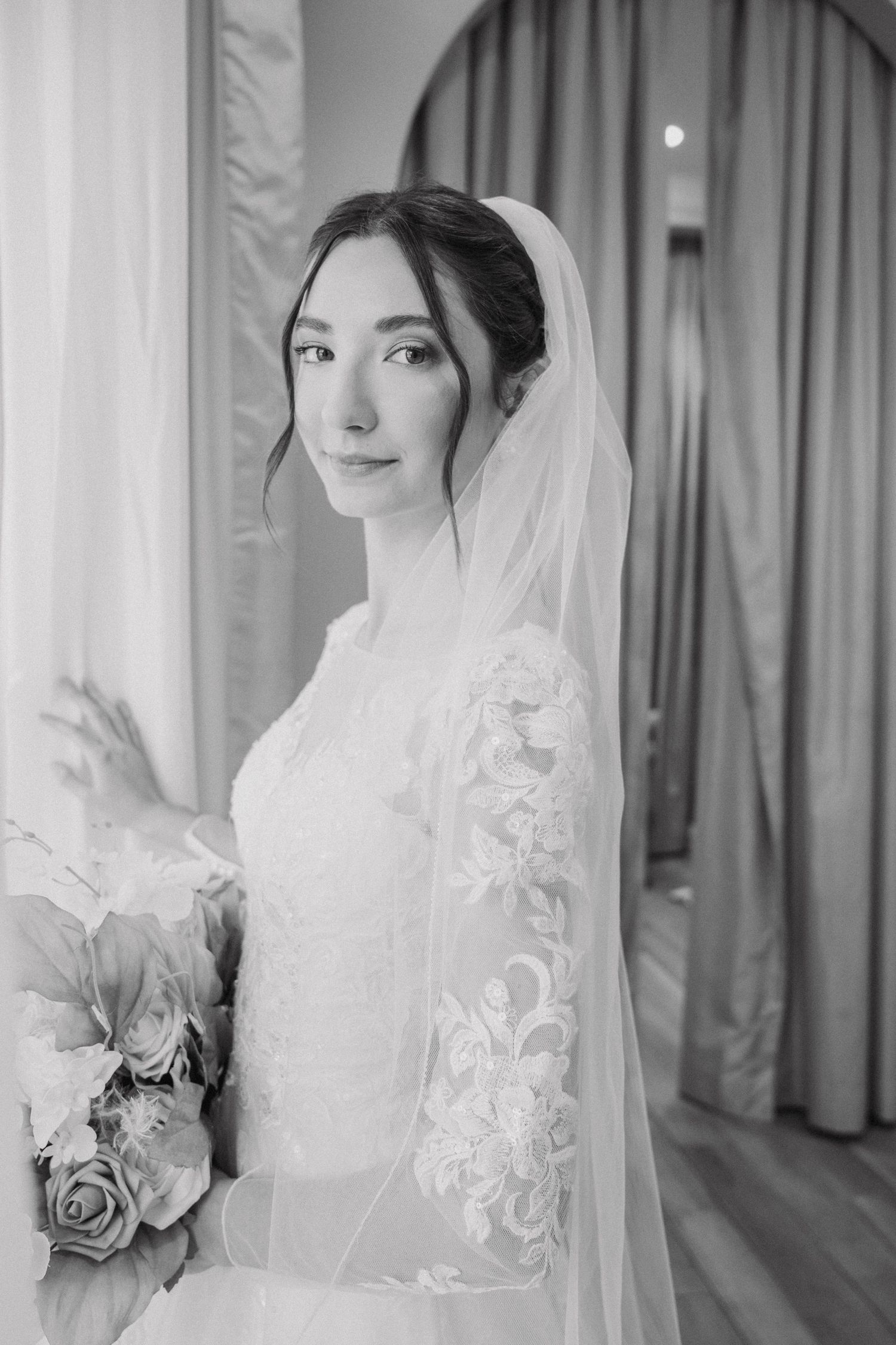 Elegant bridal portrait in white lace veil with soft lighting and curtains in background.