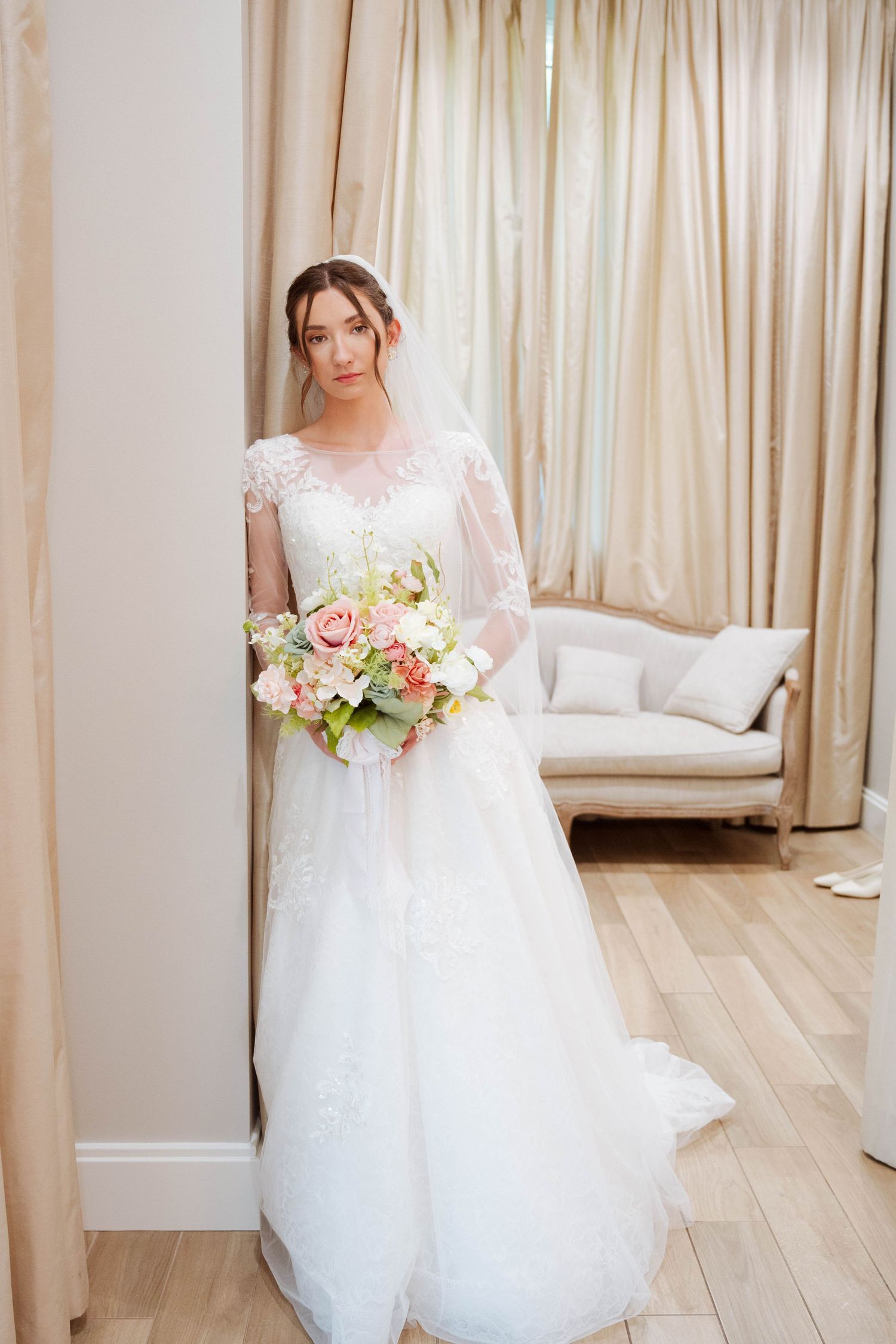 A bride in a delicate lace wedding gown holds a pink and white bouquet while standing in an elegant room.