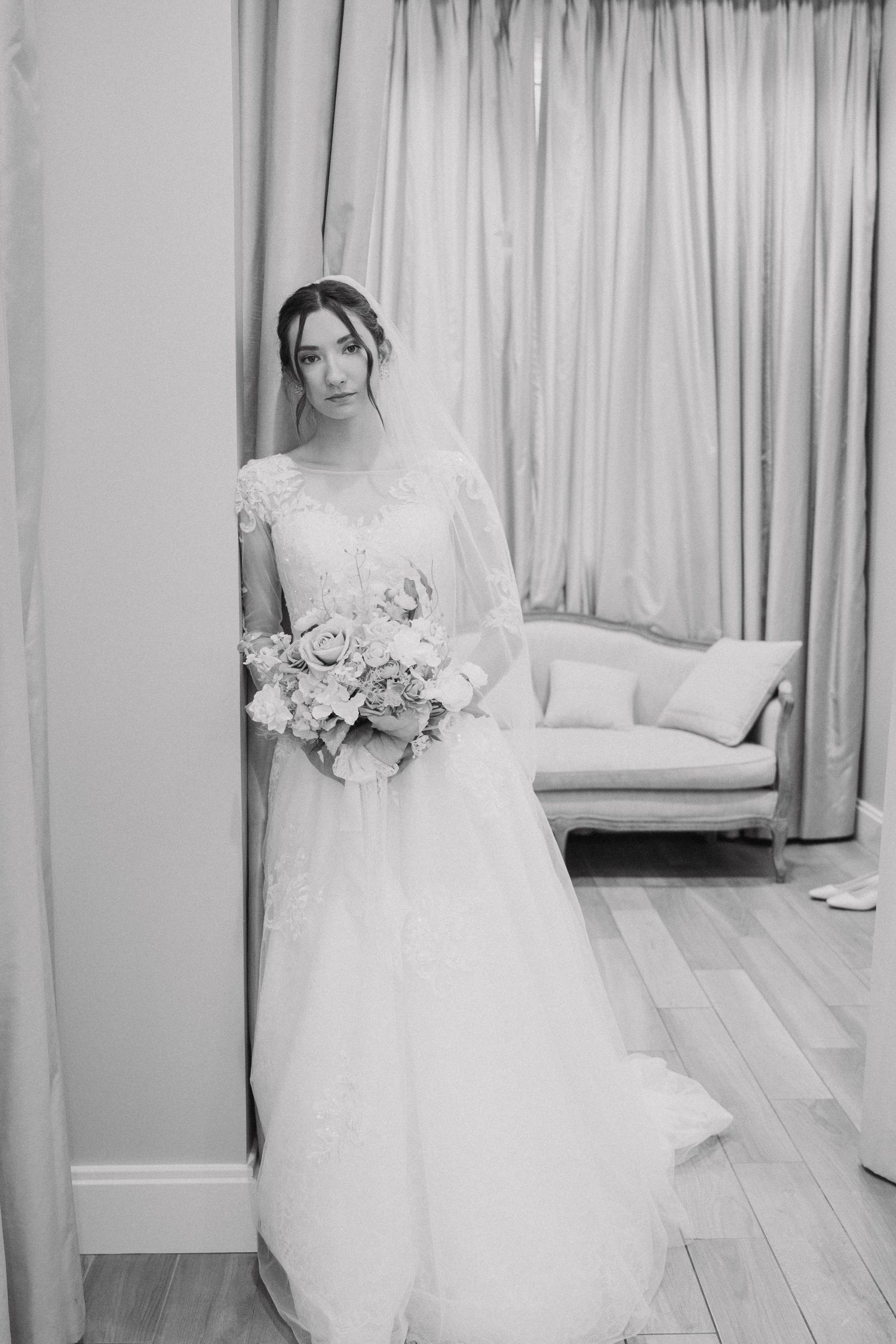 A white wedding dress with delicate lace details hangs elegantly against a light curtained background.