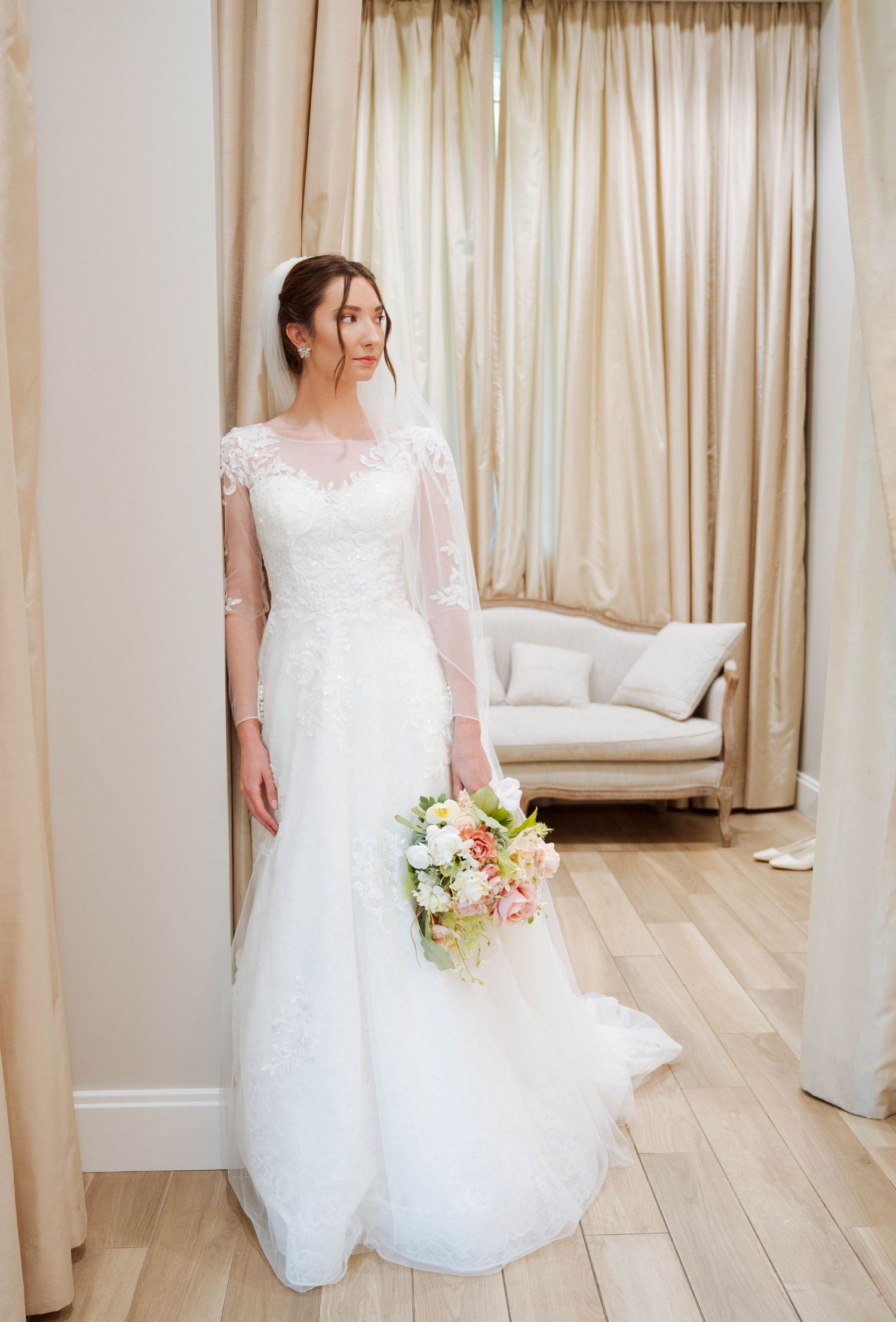 A bride in a lace wedding gown poses in a light-filled room with hardwood floors and cream curtains.