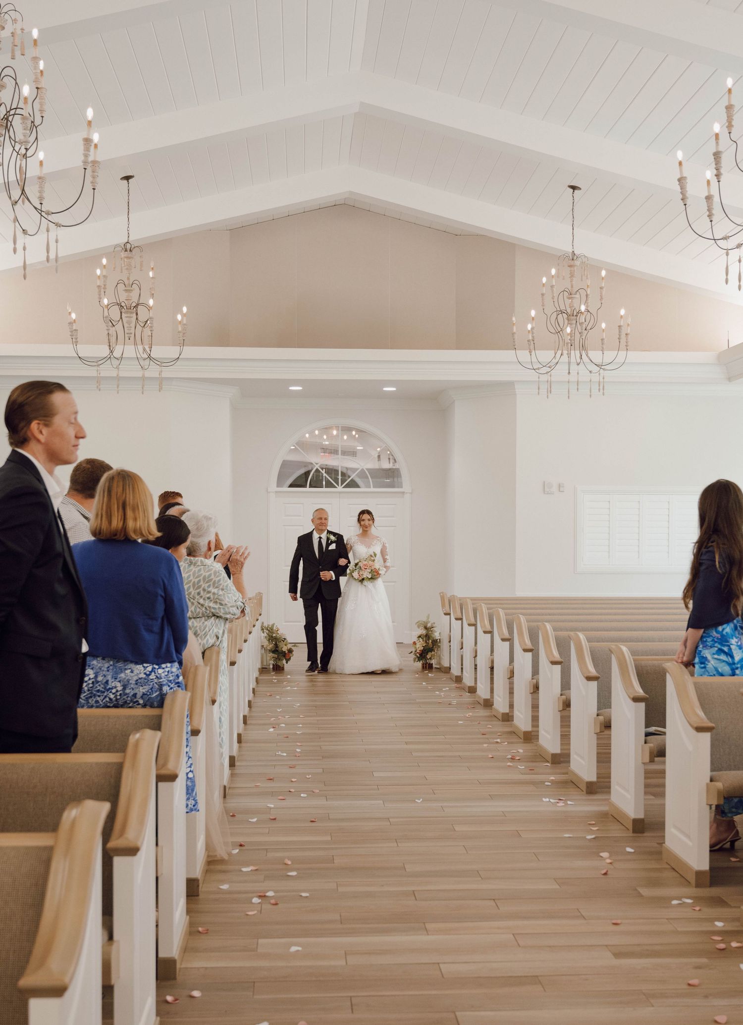 A bright chapel interior with wooden pews and crystal chandeliers during a wedding ceremony.