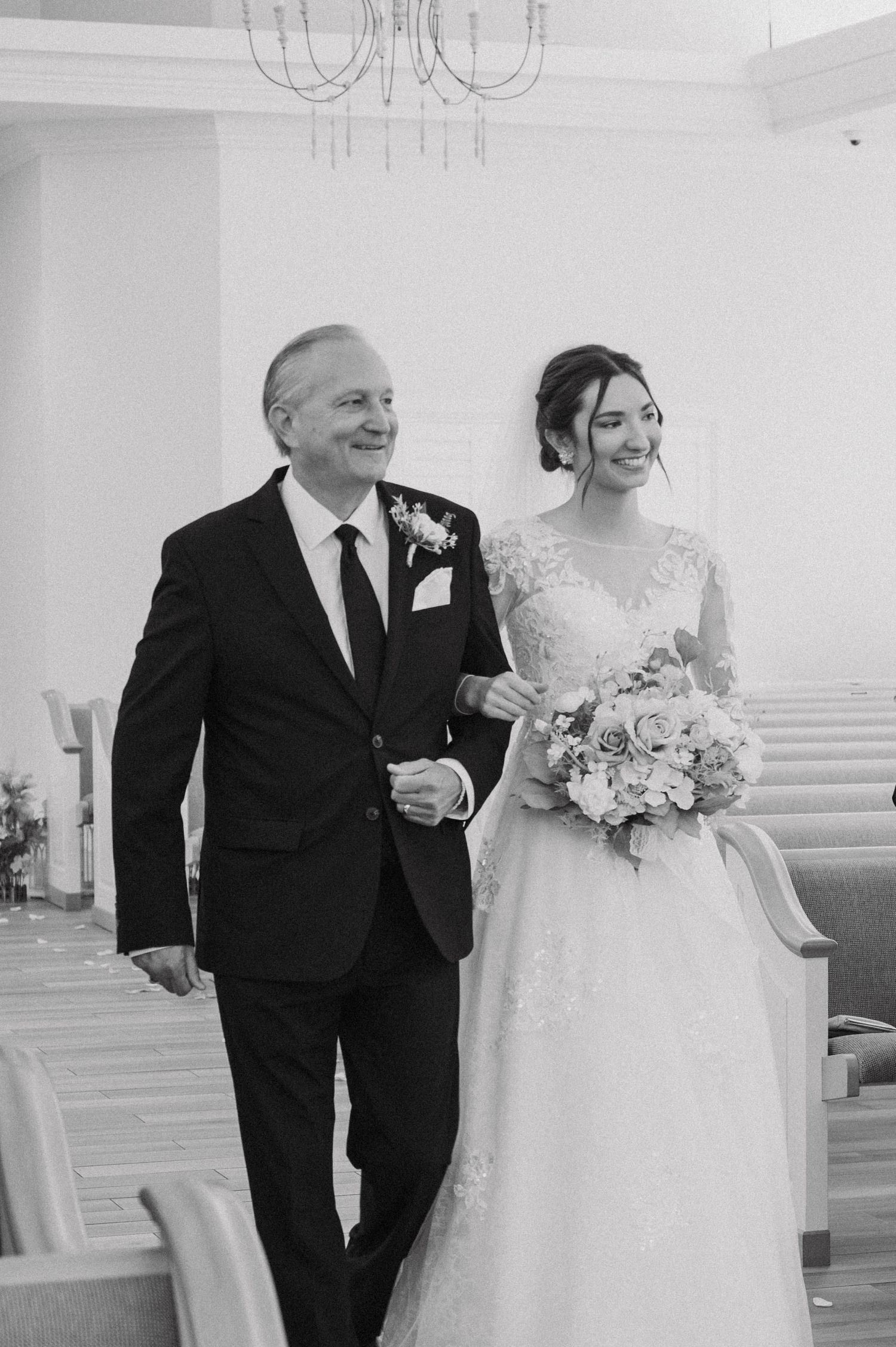 A wedding processional in a white church with a bride in lace gown and bouquet walking down the aisle.