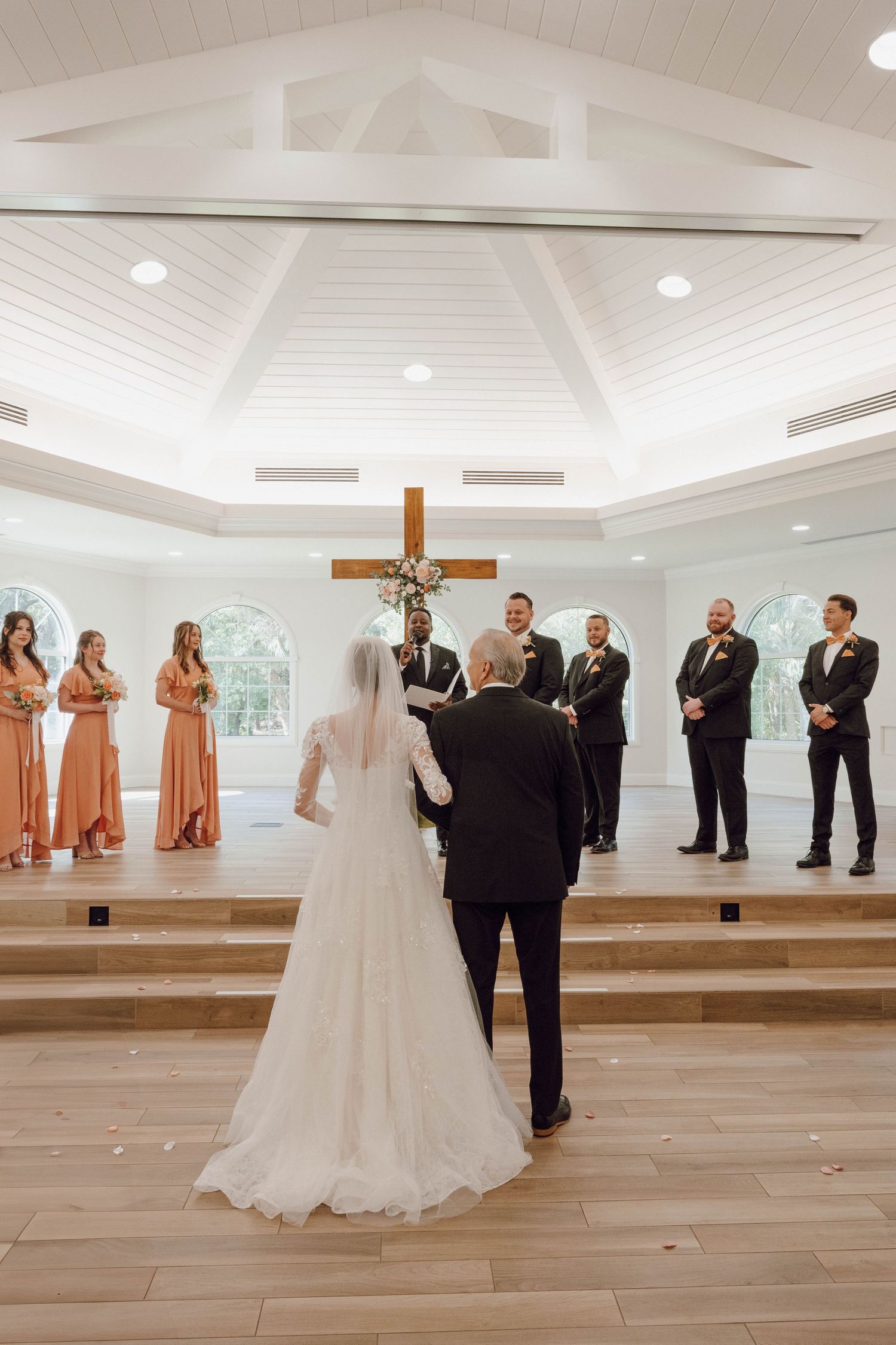 Wedding ceremony in progress inside a modern white chapel with wooden floors and vaulted ceiling.