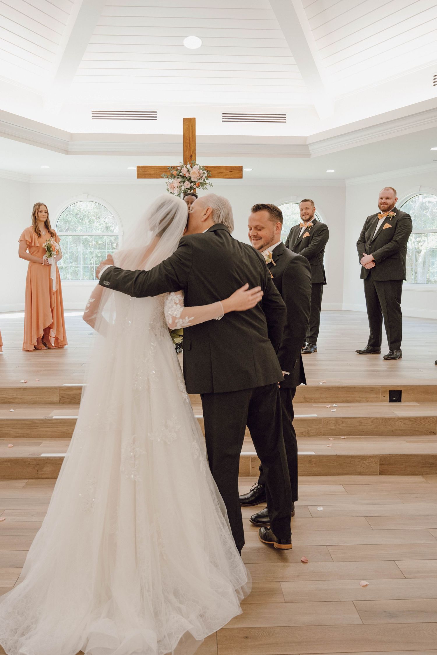 Wedding ceremony in a bright chapel with bridesmaids in peach dresses and groomsmen in black suits.