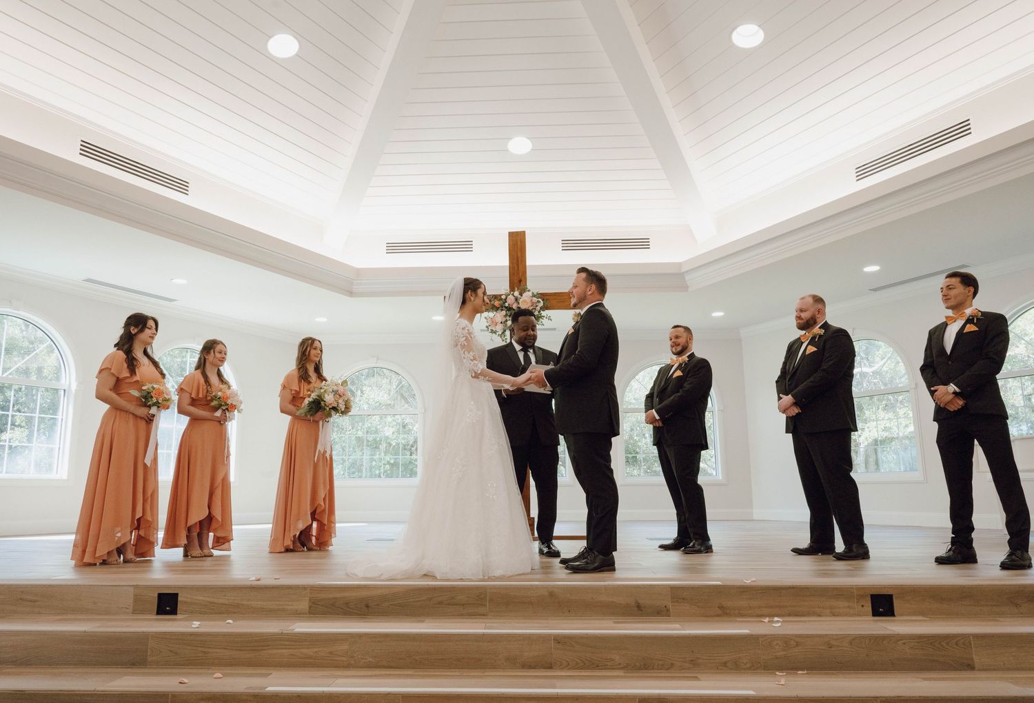 Wedding party members lined up on tiered steps in a white chapel with elegant chandeliers.