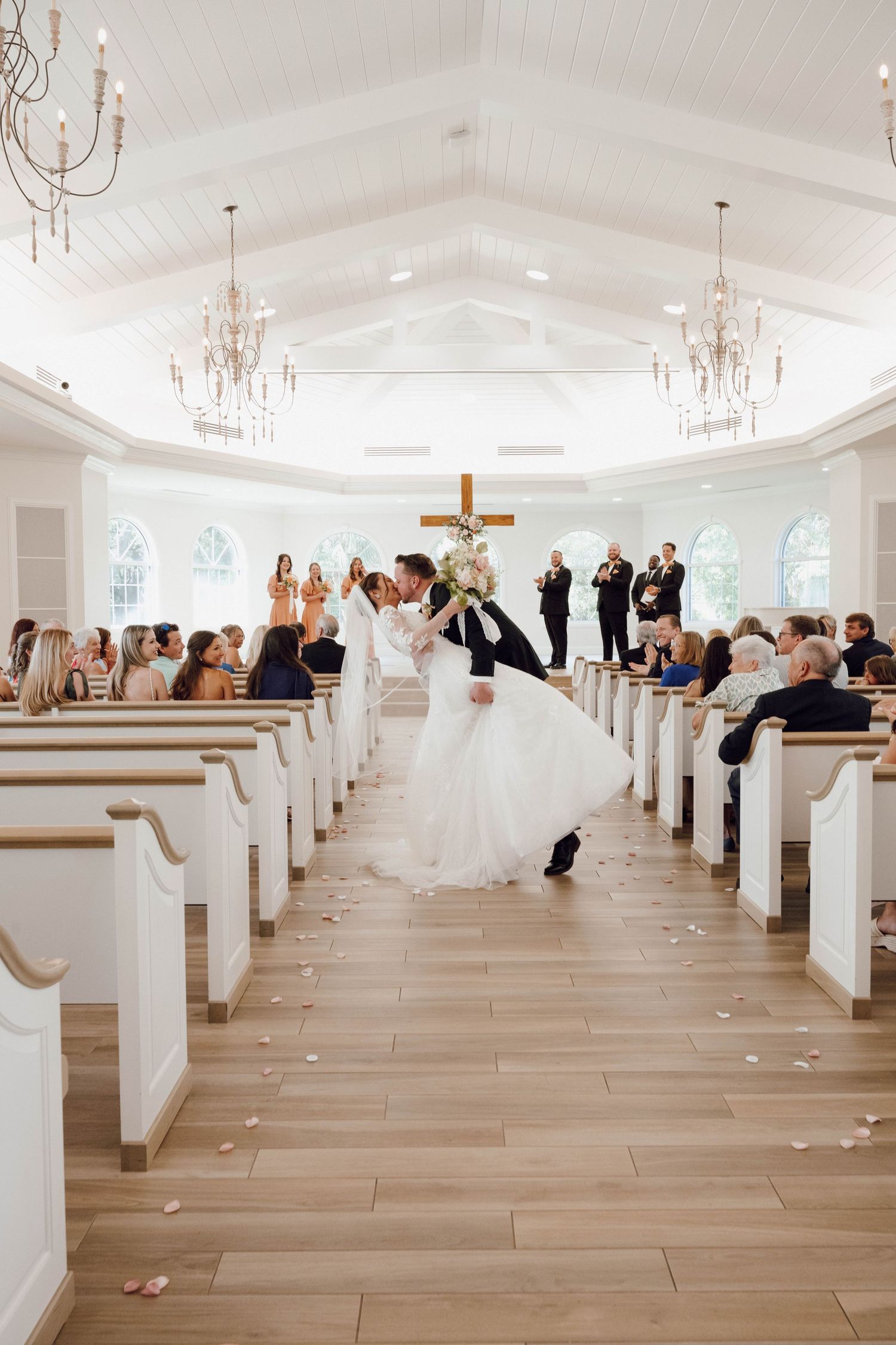 A couple shares a romantic dip during their wedding ceremony in a bright white church with crystal chandeliers.