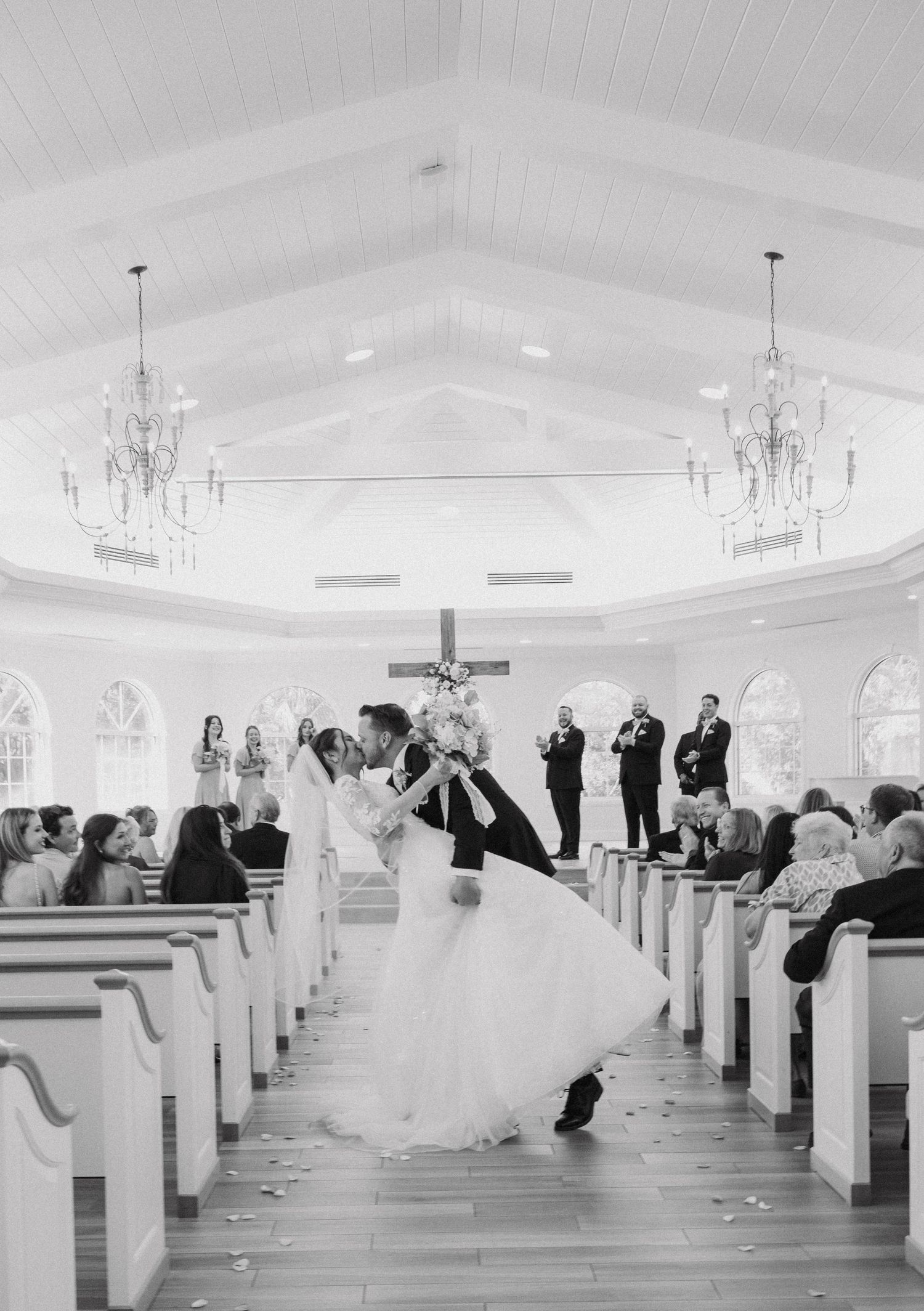 A romantic dip during a wedding ceremony in a bright white chapel with elegant chandeliers and wooden pews.