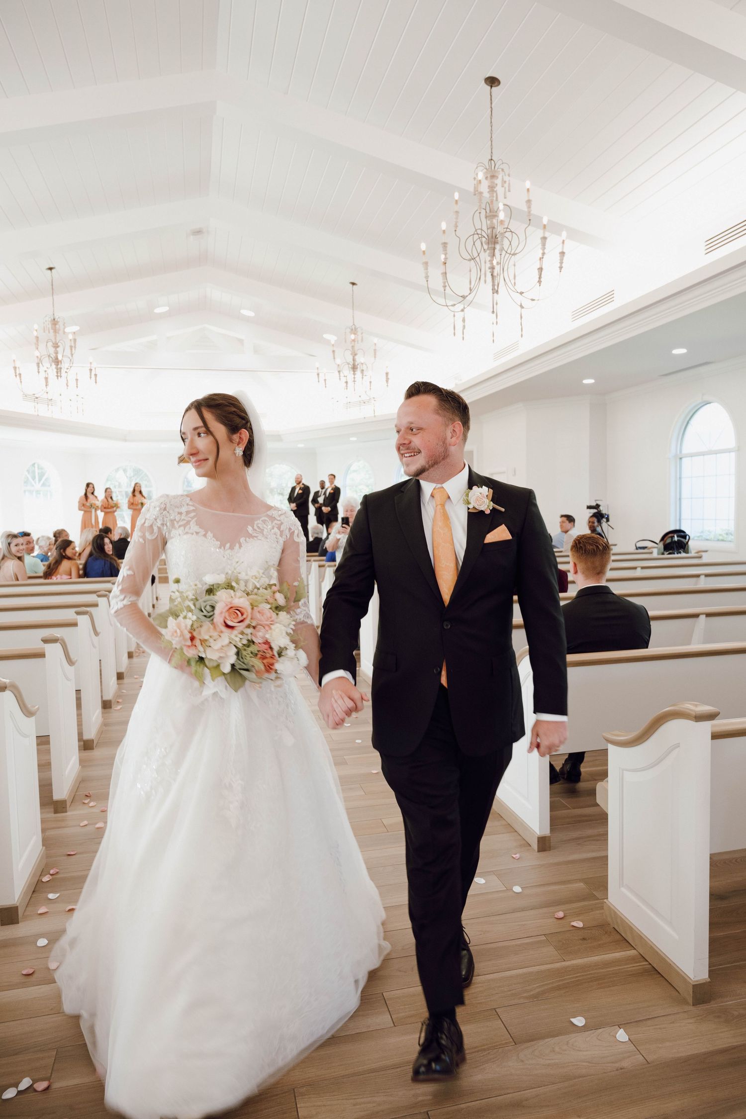 Newlyweds walk down the aisle in an elegant white chapel with crystal chandeliers and wooden pews.