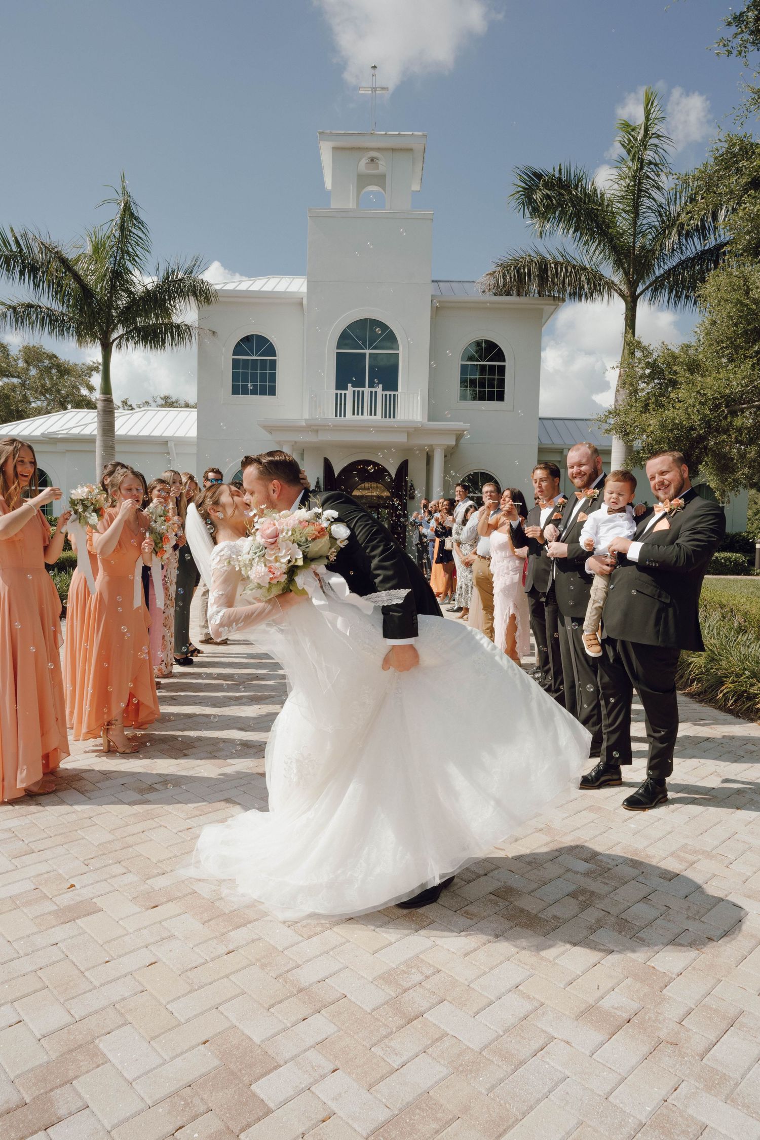 Outdoor wedding celebration with palm trees and guests in formal attire at an elegant white venue.