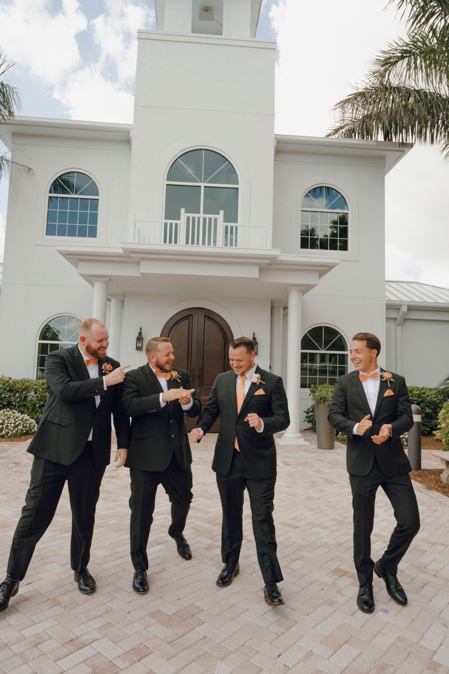 Group of groomsmen in black suits performing a synchronized dance move outside a white wedding venue.
